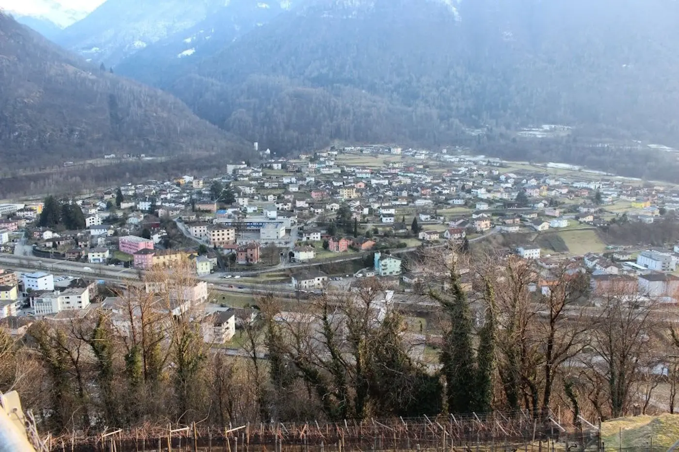 Vista panoramica di un paese montano circondato da alberi, con case colorate disposte ordinatamente in una vallata. Le montagne si ergono sullo sfondo e la luce del sole illumina il paesaggio.