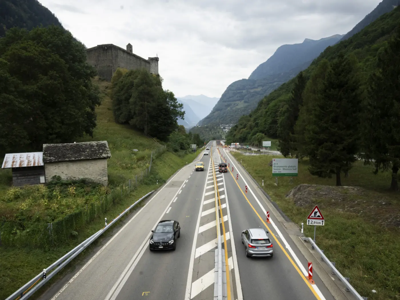Una strada a due corsie con automobili che transitano, fiancheggiata da alberi e colline. Sullo sfondo si vede un castello e montagne. Il cielo è nuvoloso.