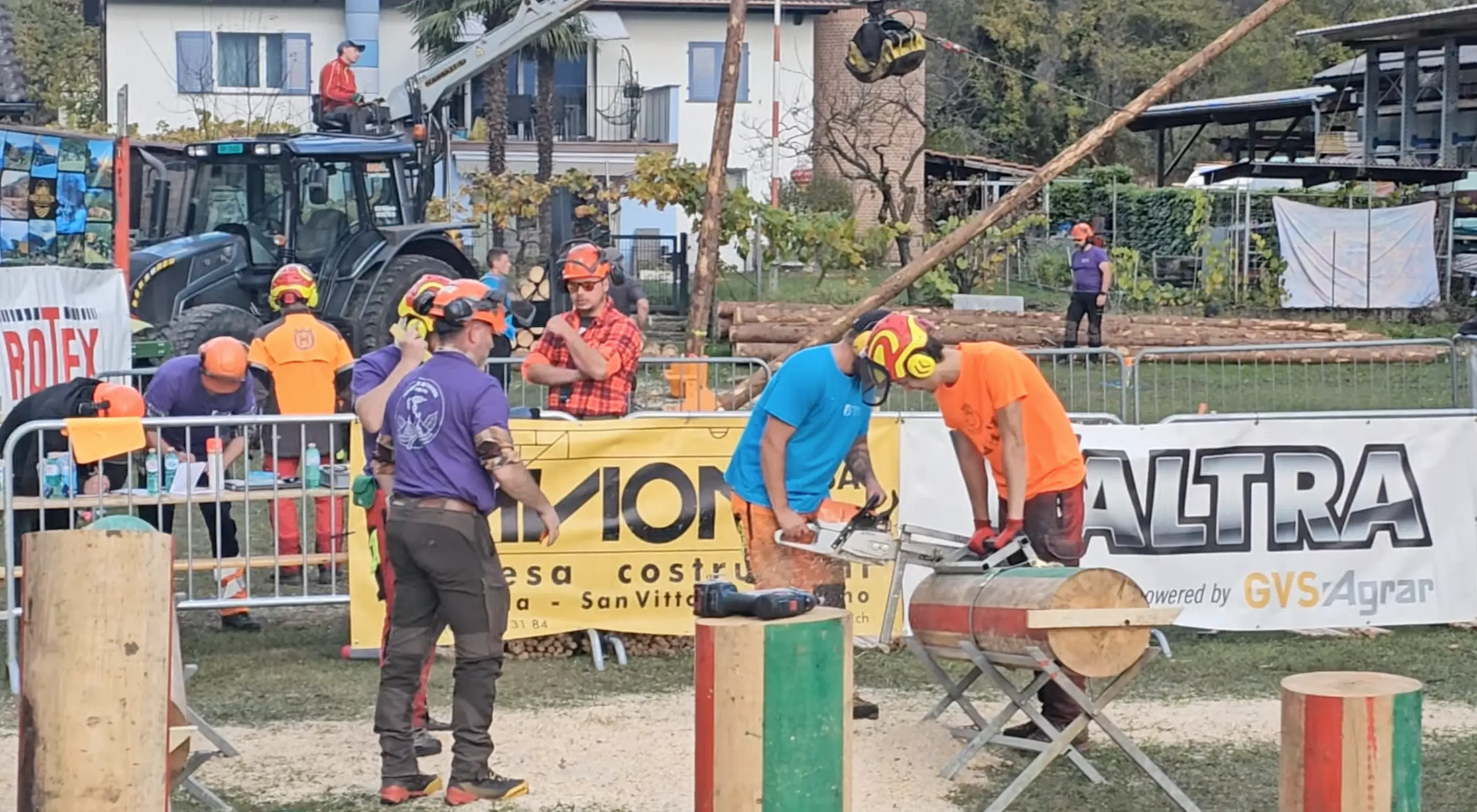 Concorrenti in un evento di abbattimento alberi, intenti a utilizzare motoseghe. Alcuni indossano caschi e abbigliamento protettivo, mentre un partecipante si prepara a tagliare un tronco. Sullo sfondo si notano att