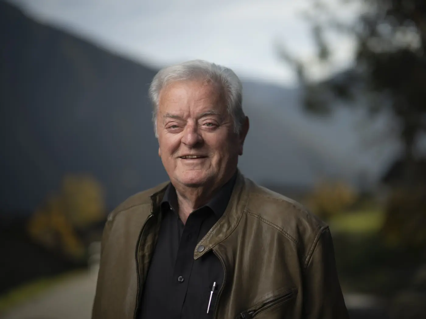 Un uomo anziano sorride davanti a un paesaggio montano, indossando una giacca di pelle e una camicia scura. Sullo sfondo si vedono alberi e montagne sfocate.