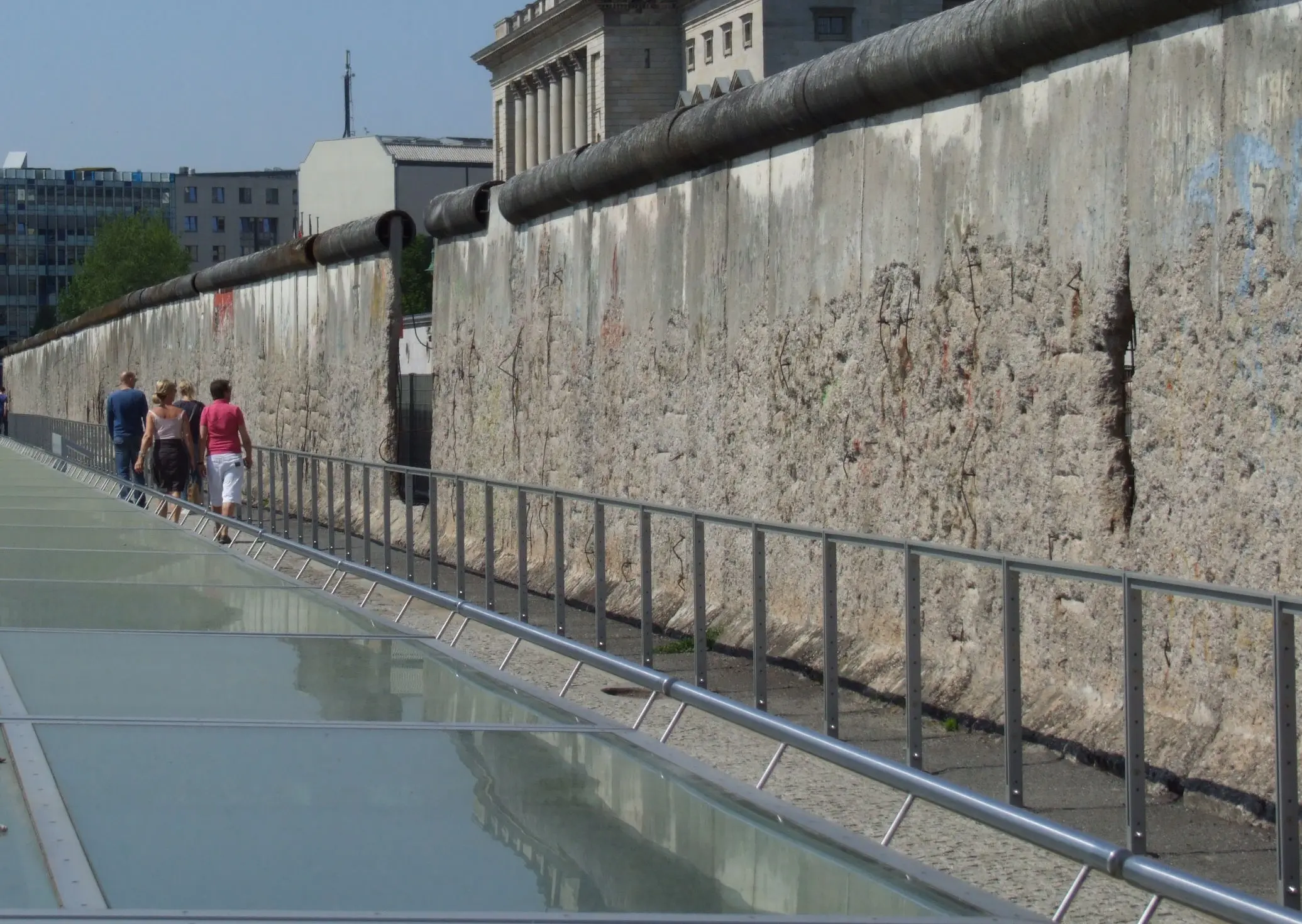 Il Muro di Berlino, con sezioni di cemento scrostatom, mentre alcuni visitatori passeggiano lungo un percorso vicino. Si intravedono edifici moderni sullo sfondo.