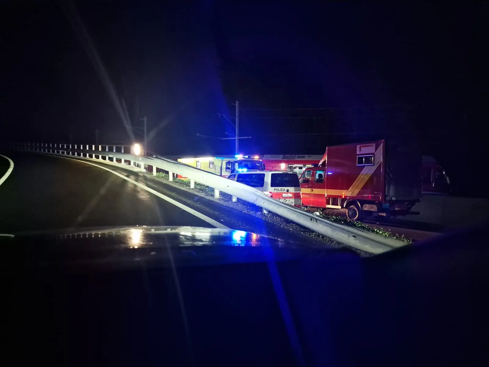 Incidente stradale notturno con veicoli di emergenza e camion lungo una strada. Le luci blu dei veicoli illuminano l'area circostante.