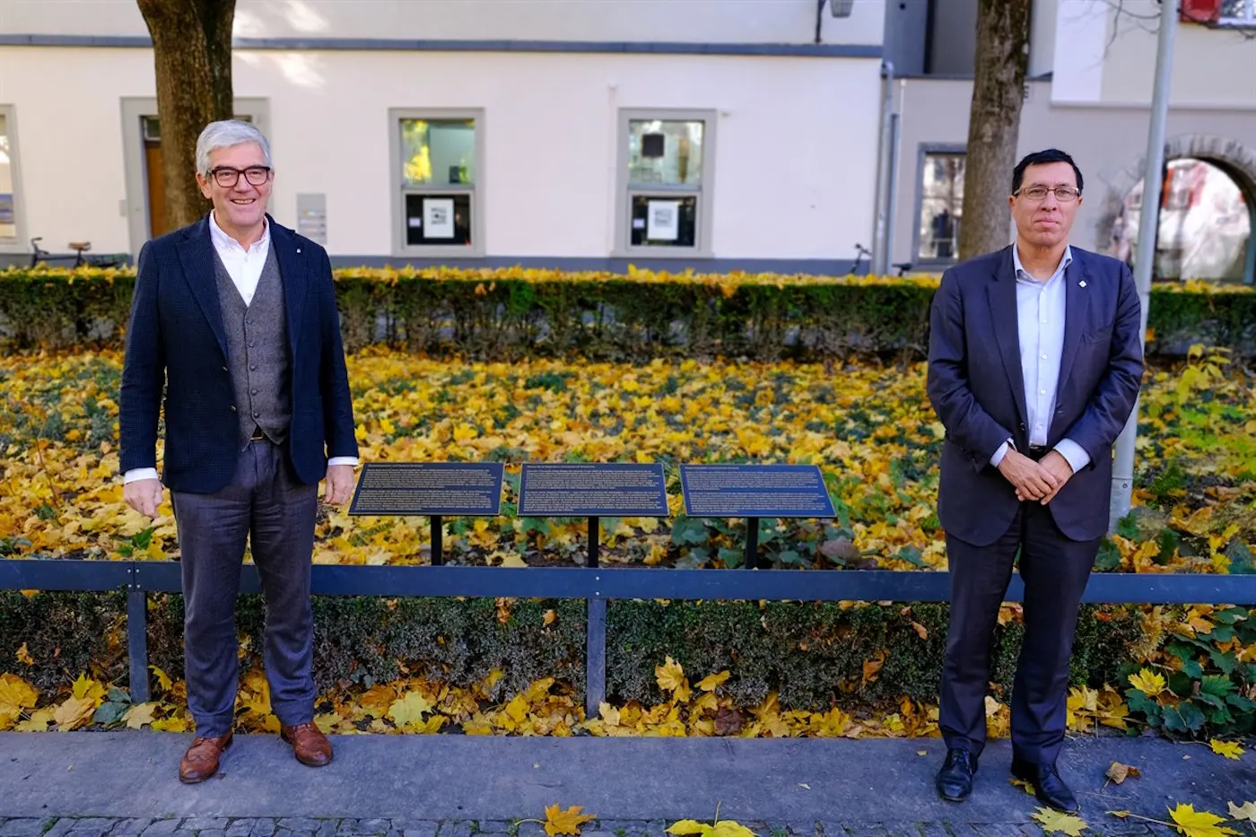 Due uomini in abiti formali posano sorridendo davanti a una panchina circondata da foglie gialle autunnali. Sullo sfondo, si vedono finestre e una parete bianca.