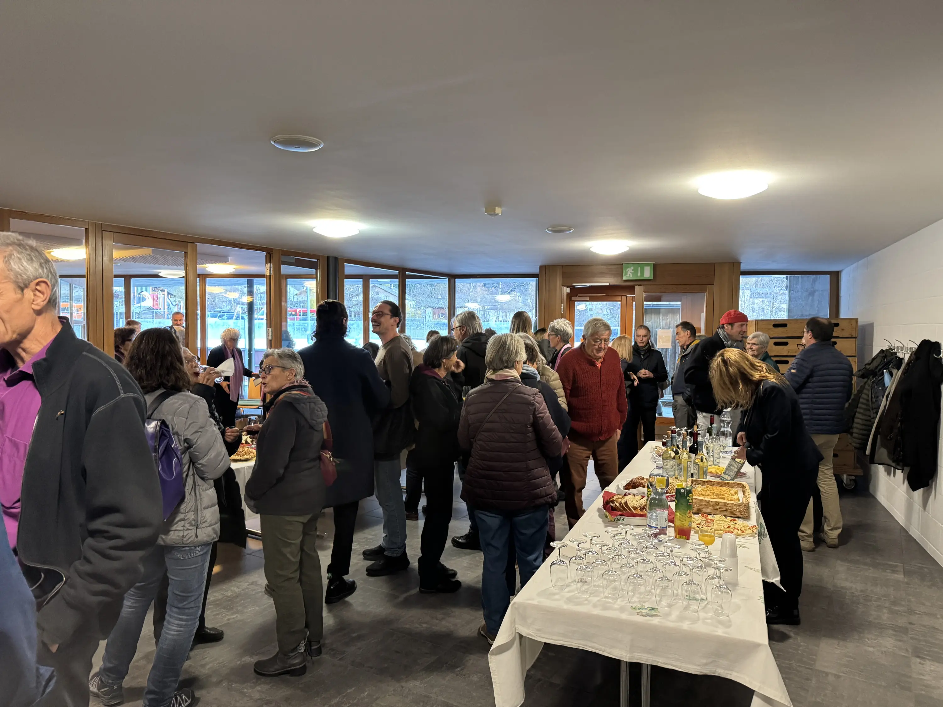 Un gruppo di persone socializza in un ambiente interno luminoso, con un buffet di cibo e bevande su un tavolo. Alcuni ospiti sono in piedi mentre altri sono seduti, creando un'atmosfera conviviale.