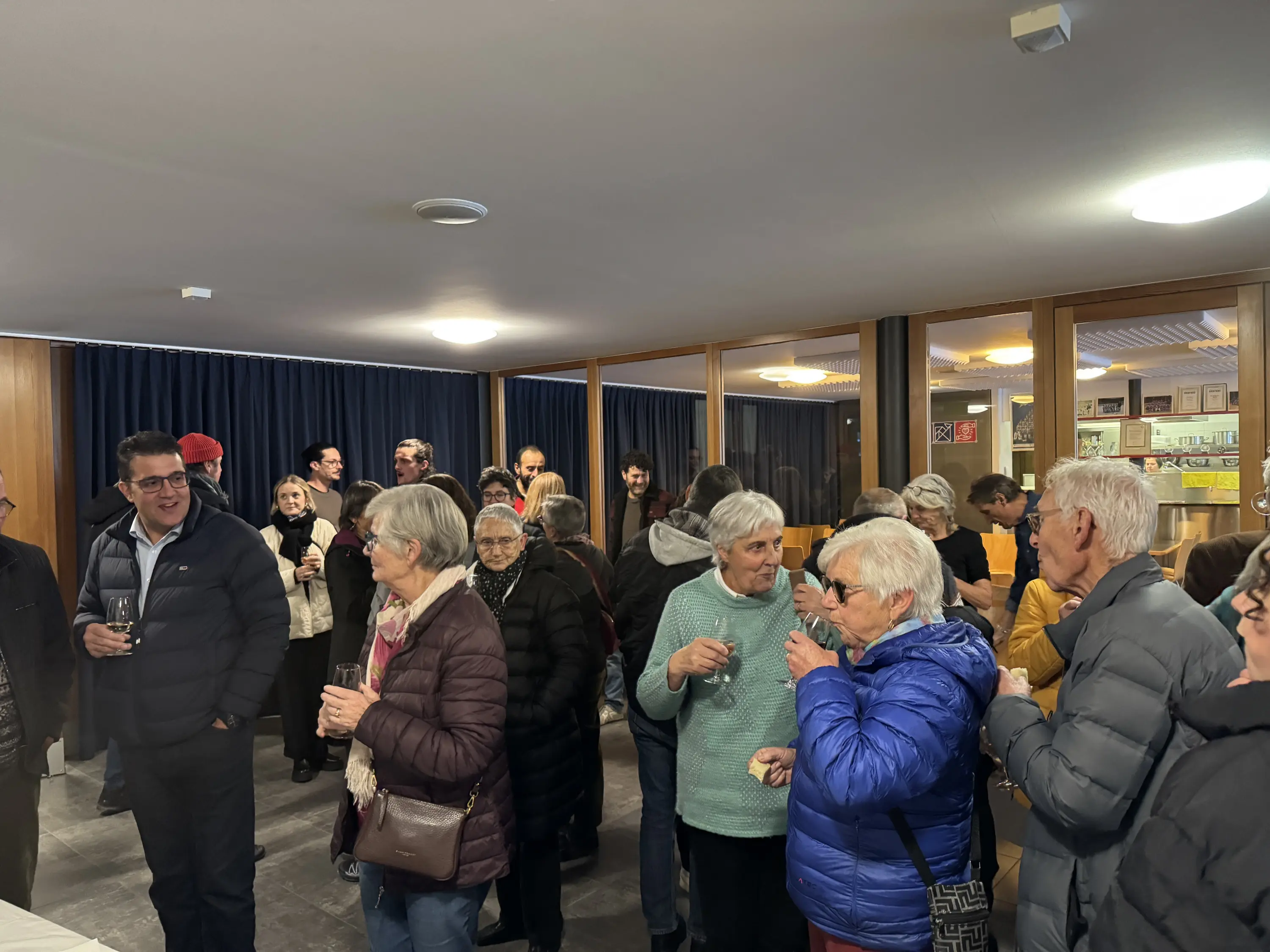 Un gruppo di persone anziane si riunisce in una sala. Alcuni sorseggiano drink e chiacchierano tra loro, mentre altri ascoltano. L'atmosfera è vivace e sociale.