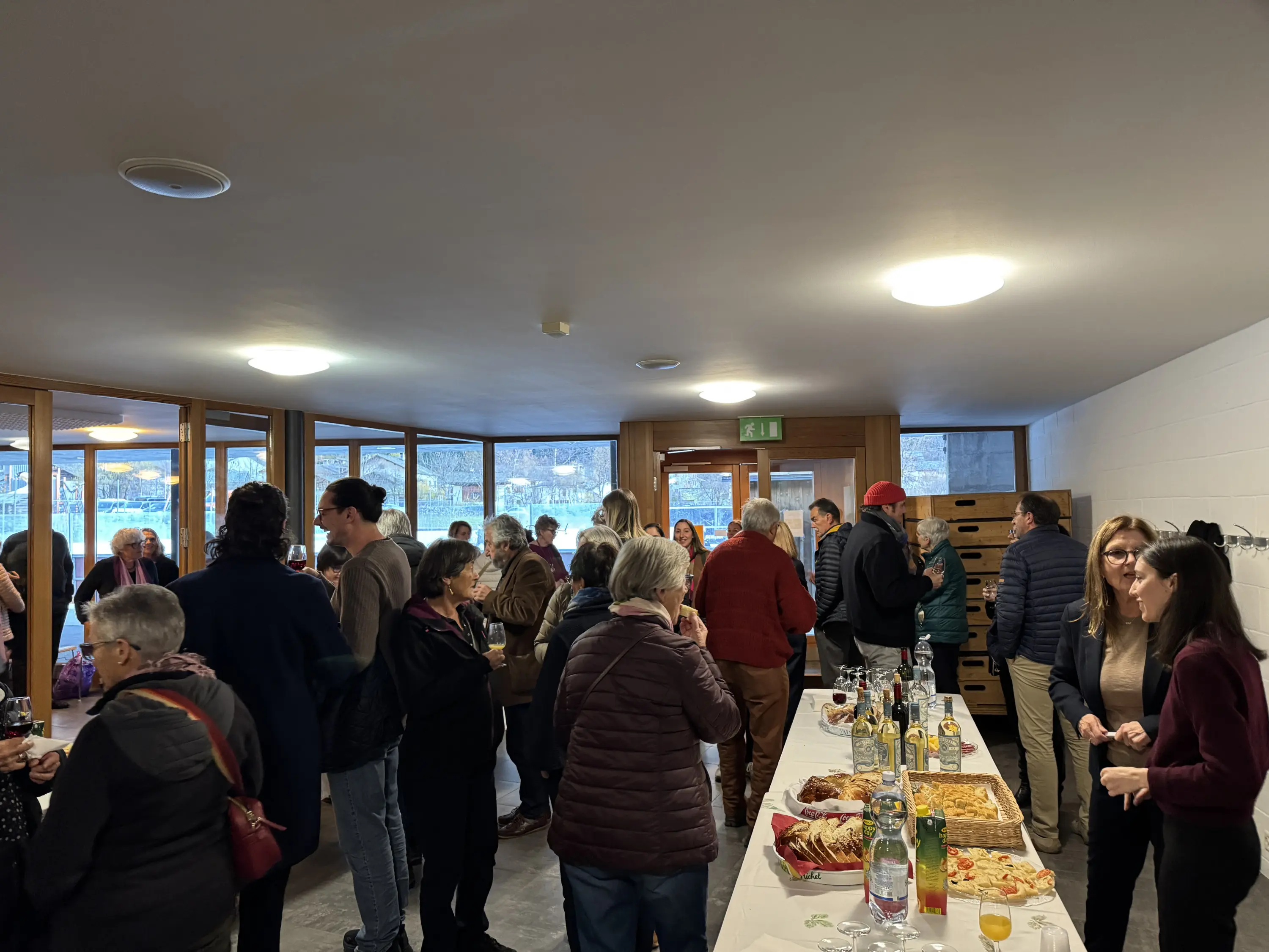 Un gruppo di persone in un ambiente interno che socializza, con tavoli imbanditi di cibo e bevande. Si possono vedere diversi ospiti conversare e gustare le pietanze. Grandi finestre lasciano entrare la luce naturale.