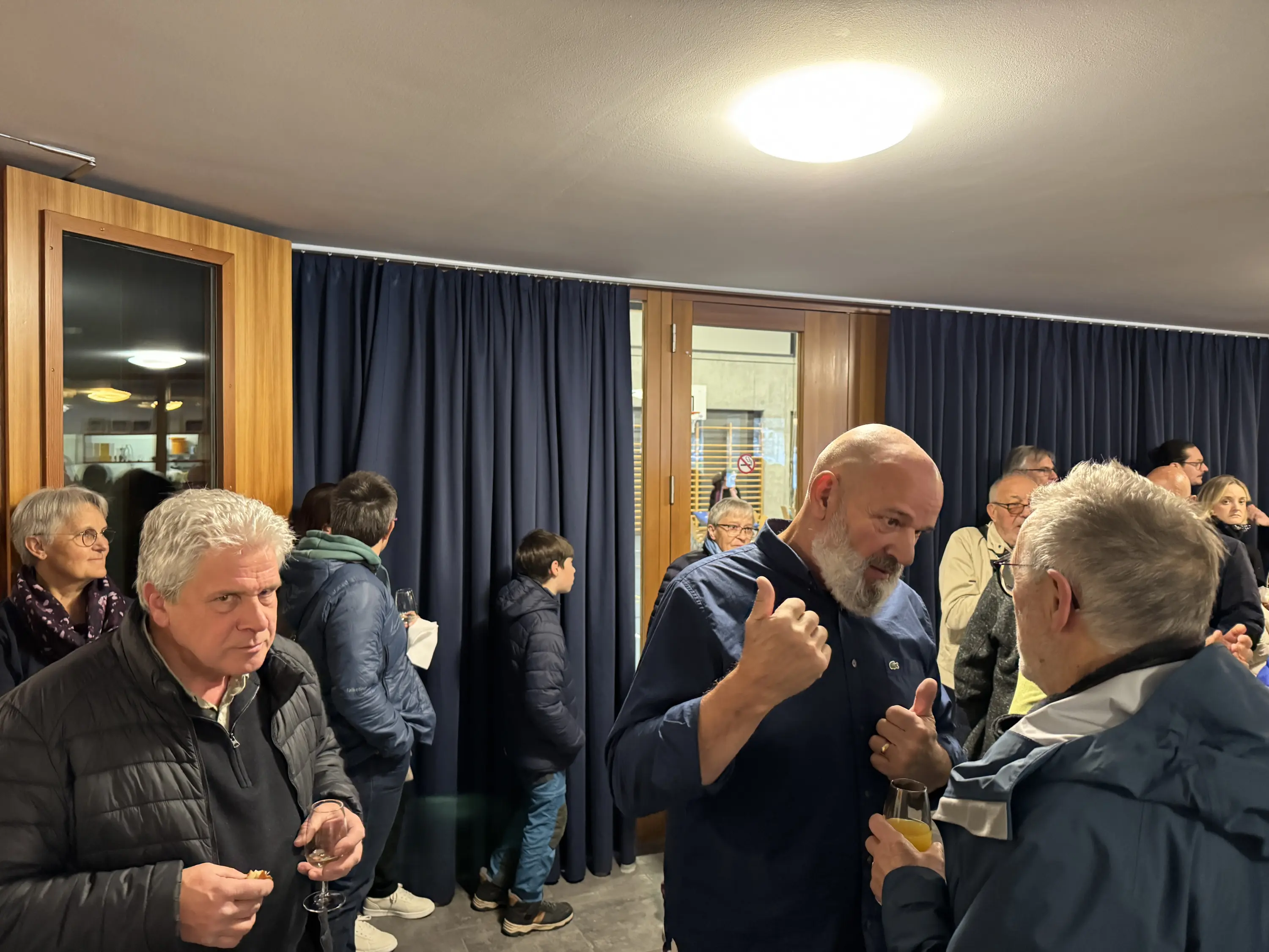 Un gruppo di persone in un ambiente interno, alcune in conversazione animata. Si vedono diverse espressioni facciali e outfit invernali. Sullo sfondo, una finestra con tende scure e altre persone che osservano o partecipano alla conversazione.