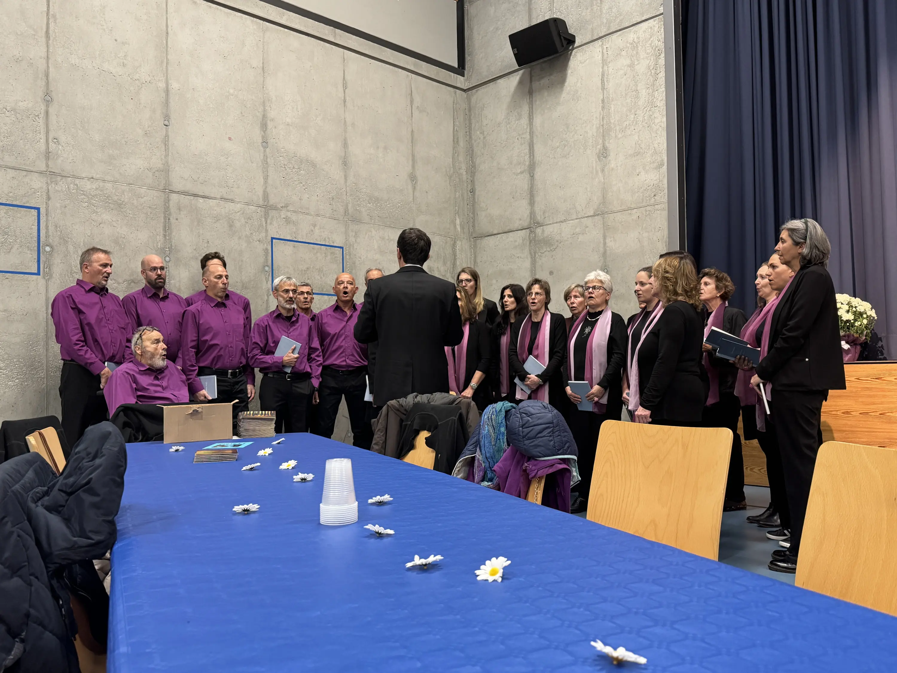 Un coro vestito con camicie viola e sciarpe rosa canta, guidato da un direttore. Il palco è in un ambiente di cemento, con un tavolo blu in primo piano decorato con fiori.