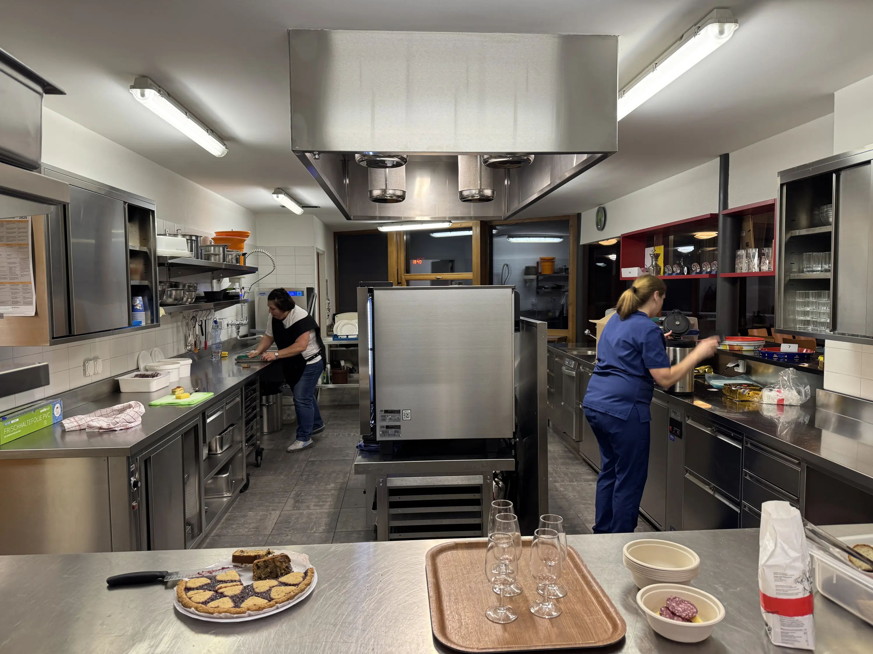 Due cuoche lavorano in una cucina professionale. Una prepara ingredienti su un piano di lavoro mentre l'altra cuoce al forno. In primo piano, vassoi con biscotti e bicchieri su un tavolo.
