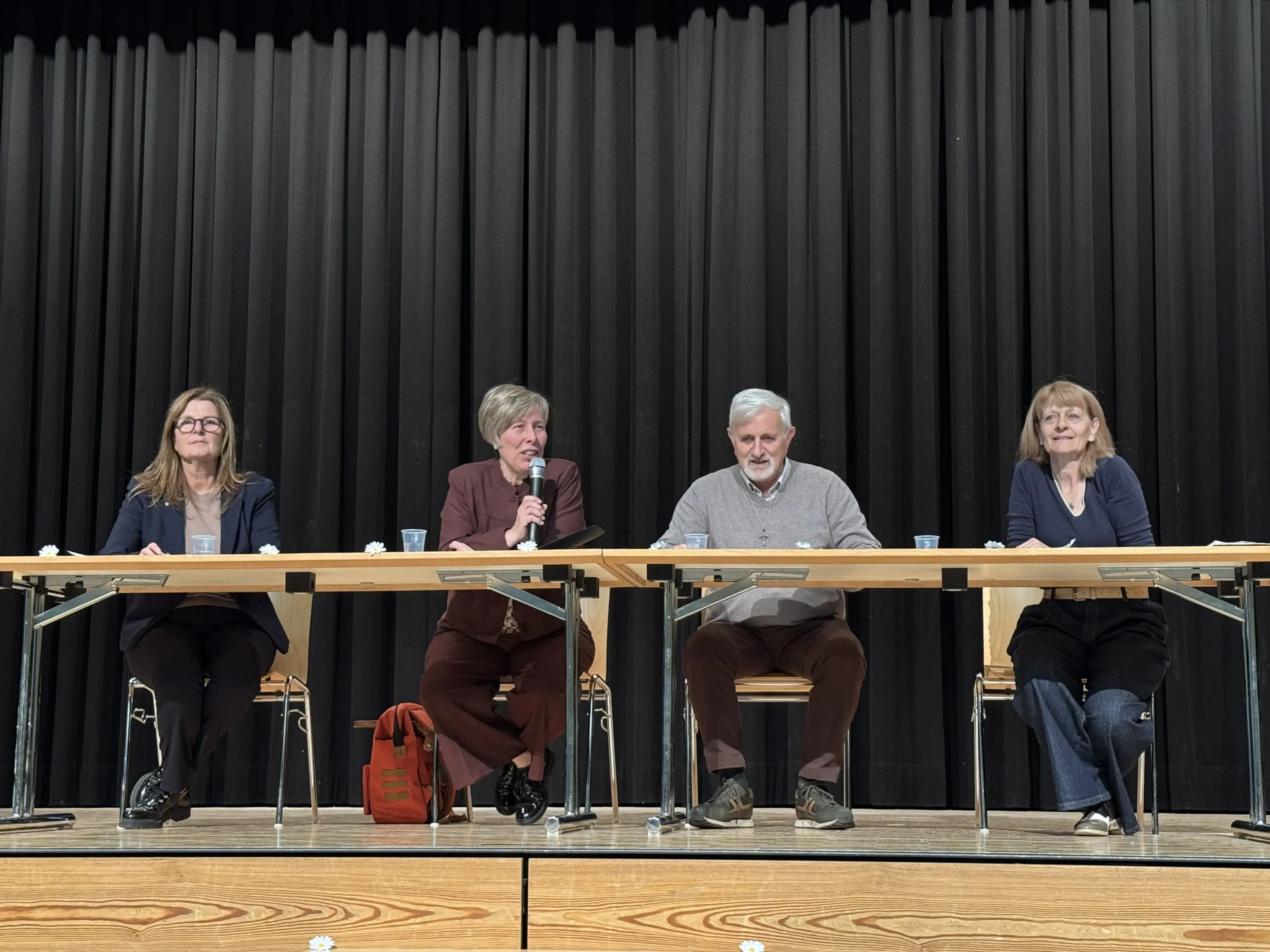Quattro relatori seduti a un tavolo su un palco. Due donne e due uomini, alcuni con microfoni, mentre discutono. Sullo sfondo, una tenda nera.
