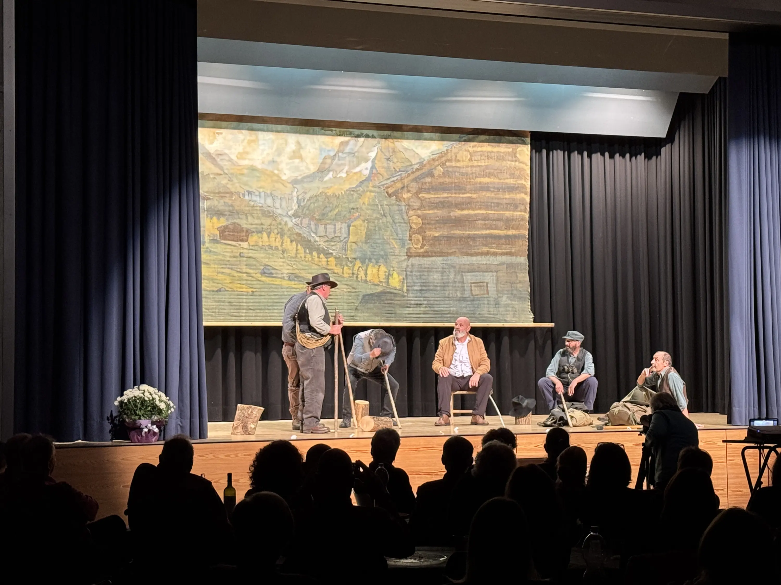 Un gruppo di attori si esibisce su un palco, con un grande sfondo di un paesaggio montano. Sono vestiti in abiti tradizionali, mentre un attore in primo piano sta interagendo con gli altri. Il pubblico è visibile in basso, in