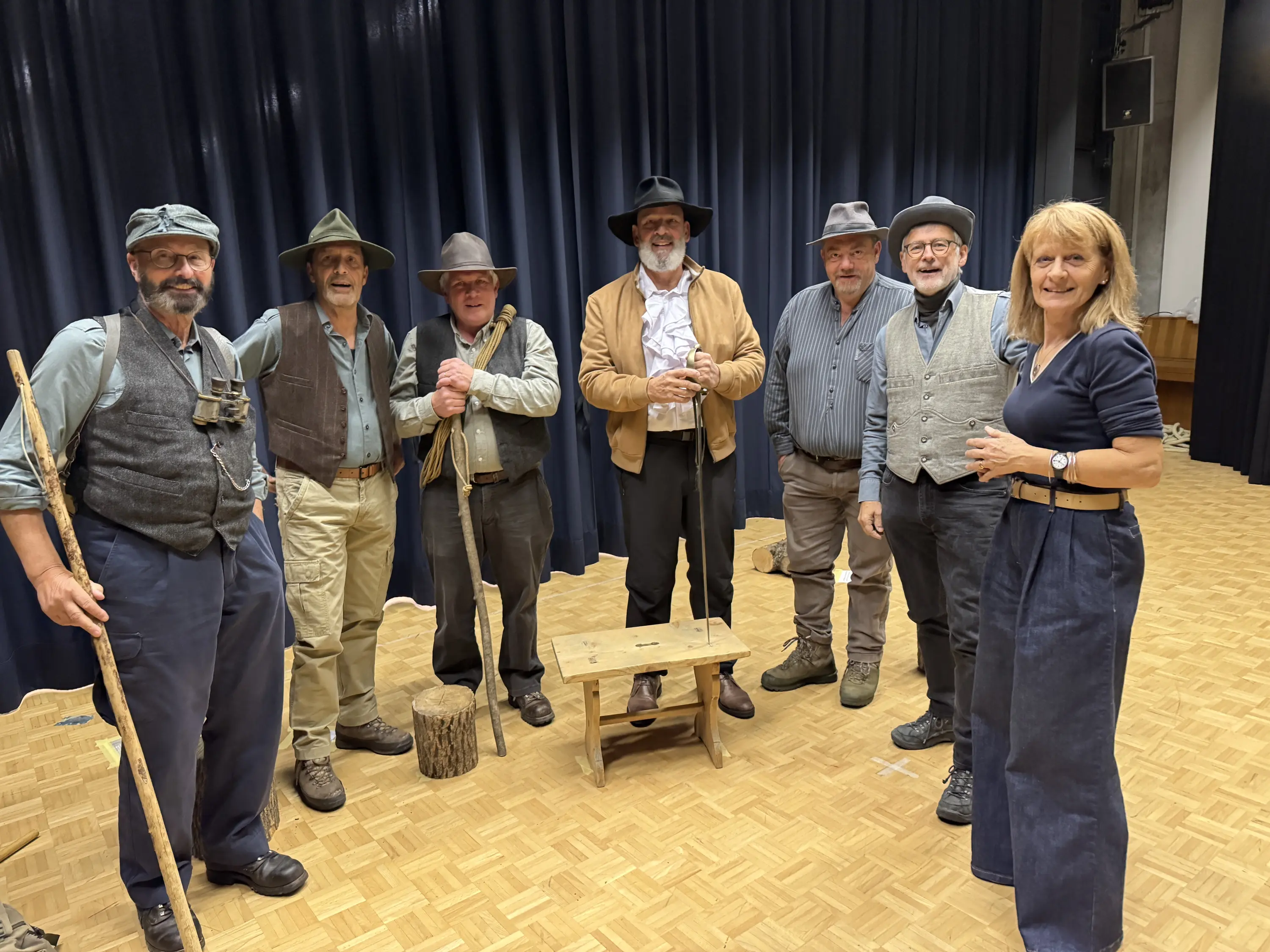 Un gruppo di otto persone in abbigliamento rustic, sorridenti e in posa, all'interno di una sala con pareti blu scuro. Ognuno tiene vari accessori come bastoni e cappelli. Un tavolino di legno è situato al centro.