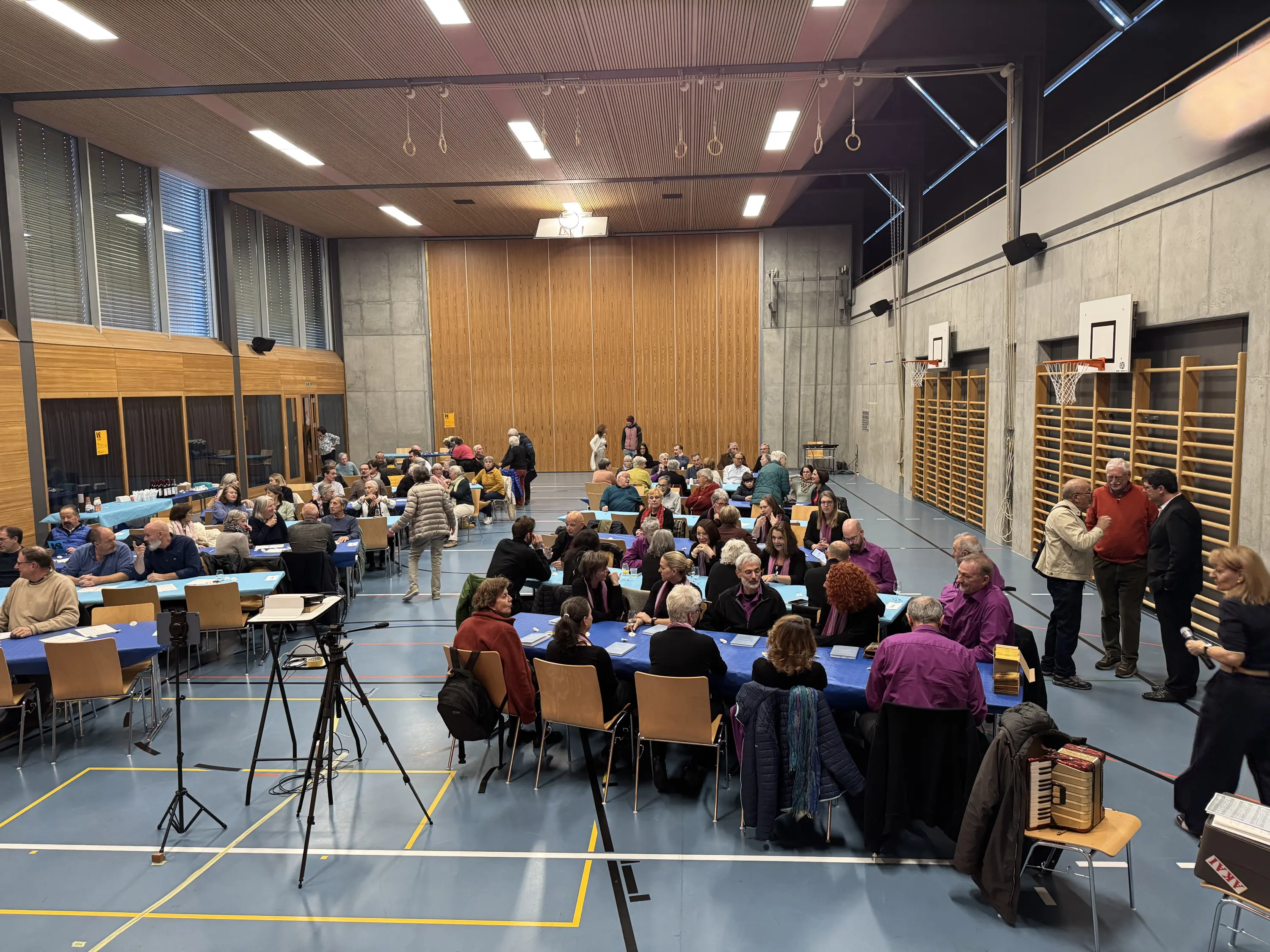 Una grande sala con tavoli blu disposti per diverse persone sedute. Molti partecipanti interagiscono fra loro, mentre alcuni si concentrano su attività diverse. L'ambiente appare luminoso e moderno, con pareti in legno e ampie finestre.