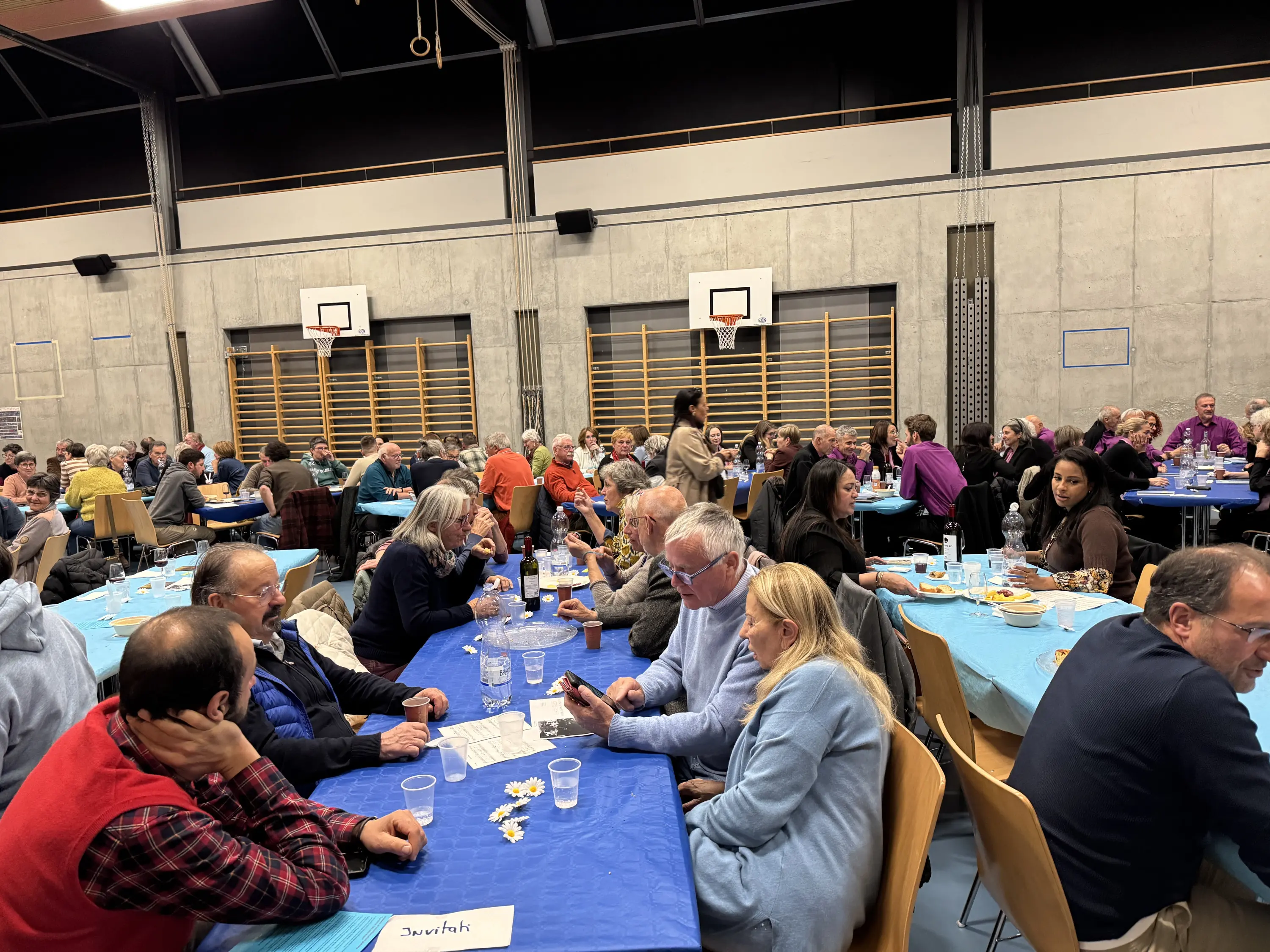 Una sala affollata con molte persone sedute a tavoli coperti di tovaglie azzurre. I partecipanti sono impegnati in conversazioni, mentre un gruppo è in piedi sullo sfondo. Il clima è vivace e informale.