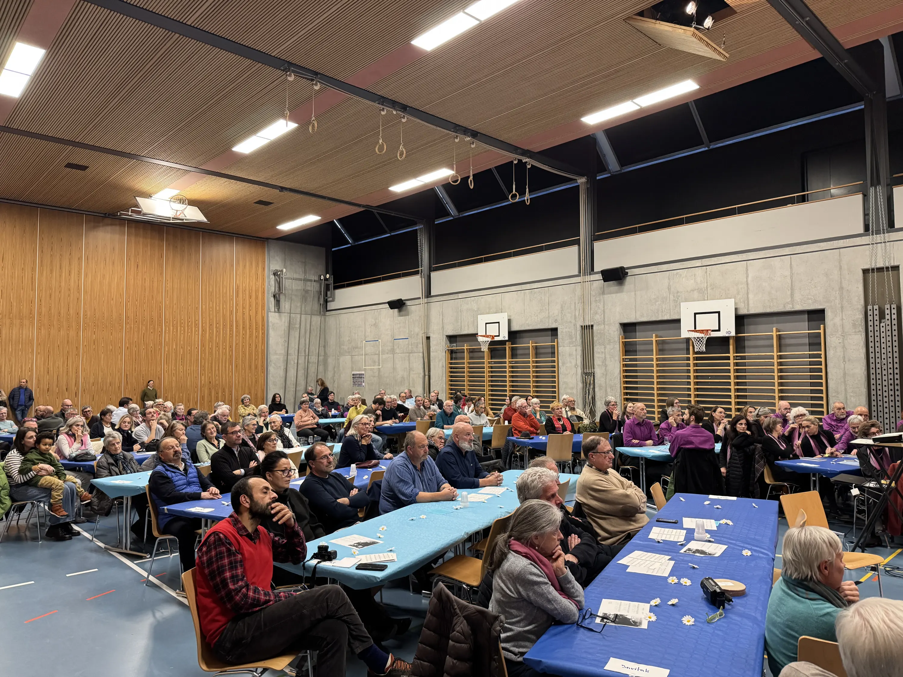 Una sala grande piena di persone anziane sedute a tavoli con tovaglie azzurre. Alcuni partecipanti ascoltano attentamente, mentre altri chiacchierano tra loro. Sullo sfondo ci sono pareti con attrezzature sportive e porte.