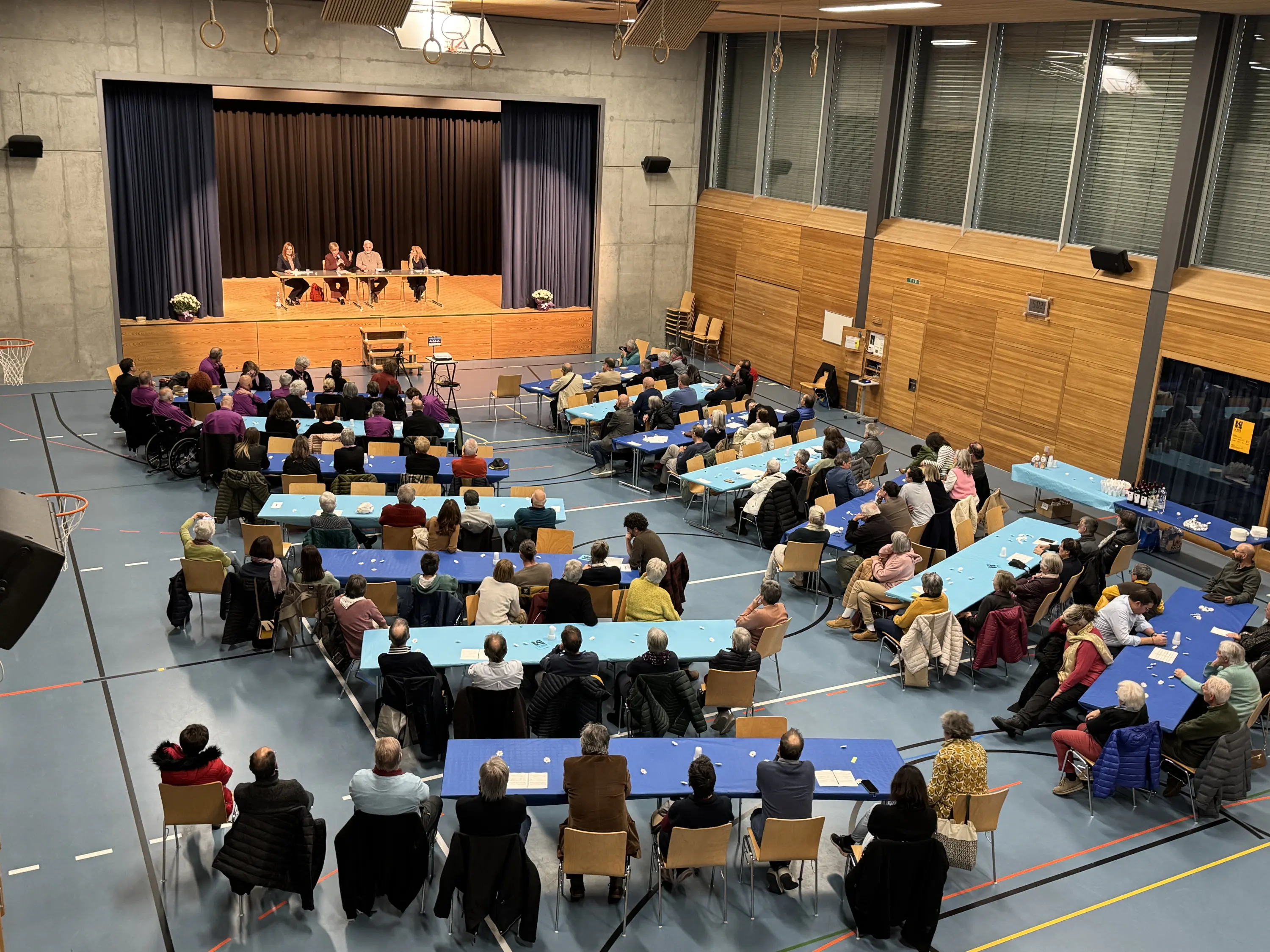 Un auditorium affollato durante un evento, con un gruppo di relatori sul palco e il pubblico seduto di fronte su diverse file di sedie. Tavoli blu sono disposti in modo ordinato e alcune piante decorative sono visibili sul palco.
