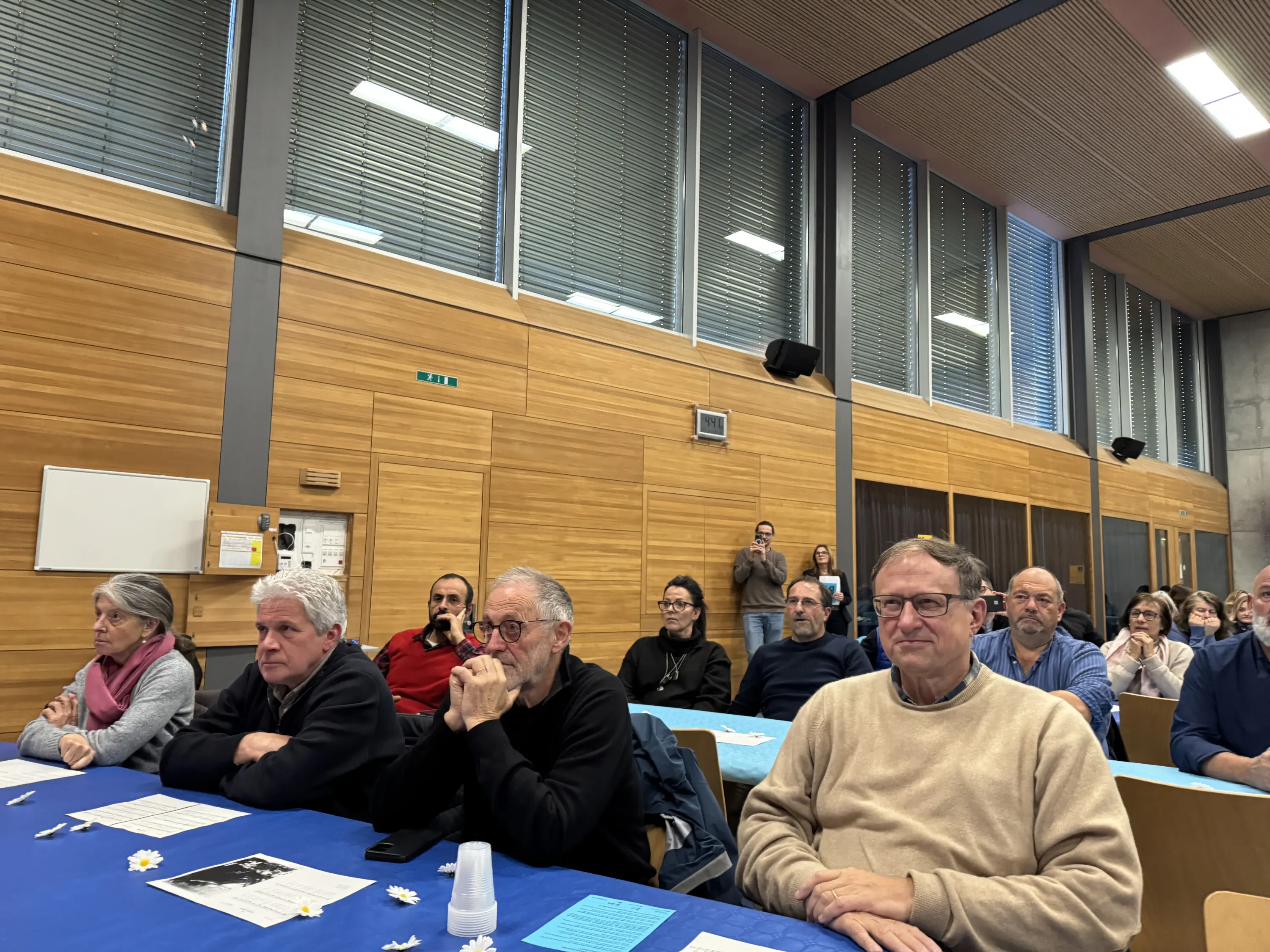 Un gruppo di persone sedute in un'aula con tavoli coperti da un tovagliolo blu. Sono attentamente concentrate sulla presentazione, con diversi volti visibili e attitudini varie. Alcuni hanno espressioni pensierose, mentre altri sono più rilassati.