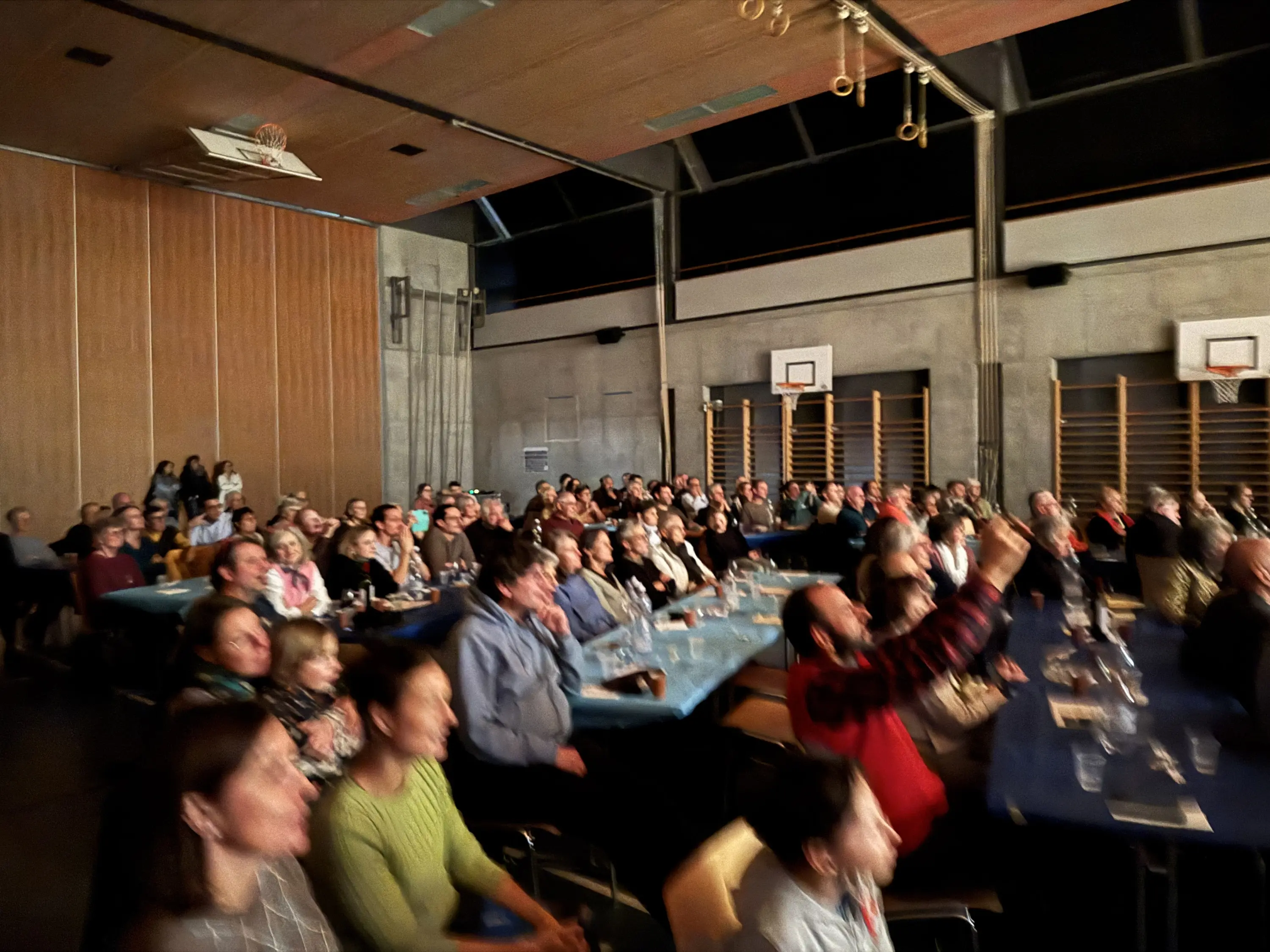 Una sala affollata con un pubblico vario che assiste a un evento, con tavoli blu e una parete di legno sullo sfondo. Molti volti sorridenti e incuriositi, alcuni che applaudono o scattano foto.