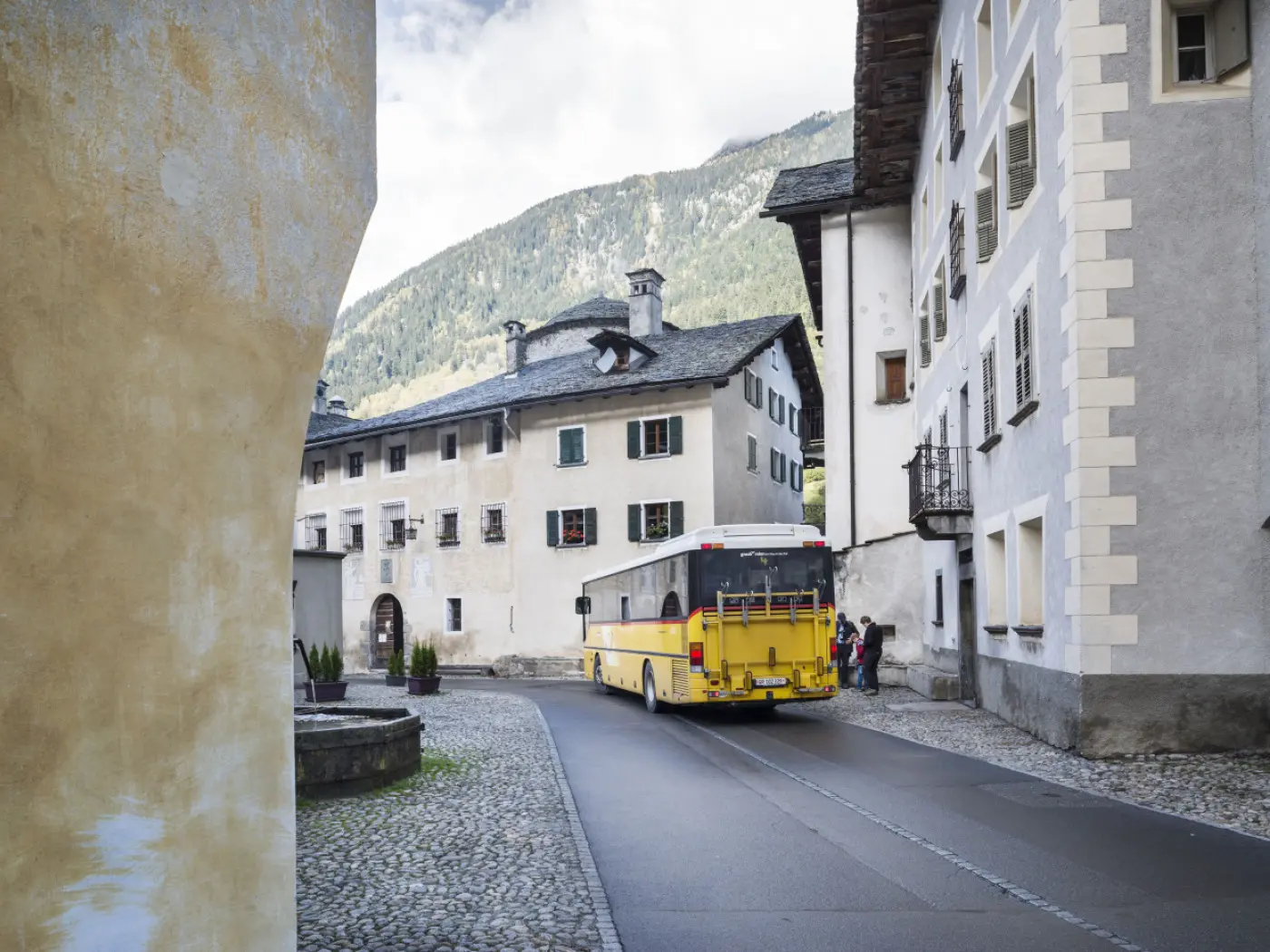 Un autobus giallo gira l'angolo di un vicolo, circondato da edifici storici con finestre colorate e montagne sullo sfondo.