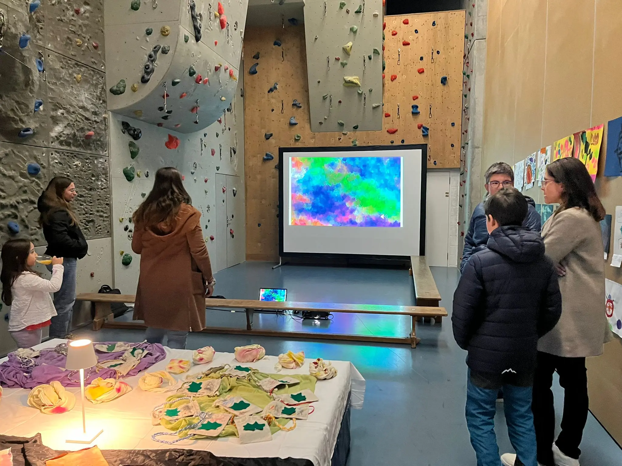 Una sala con un muro per l'arrampicata e una proiezione colorata sullo schermo. Diverse persone si riuniscono attorno a un tavolo con oggetti decorativi.