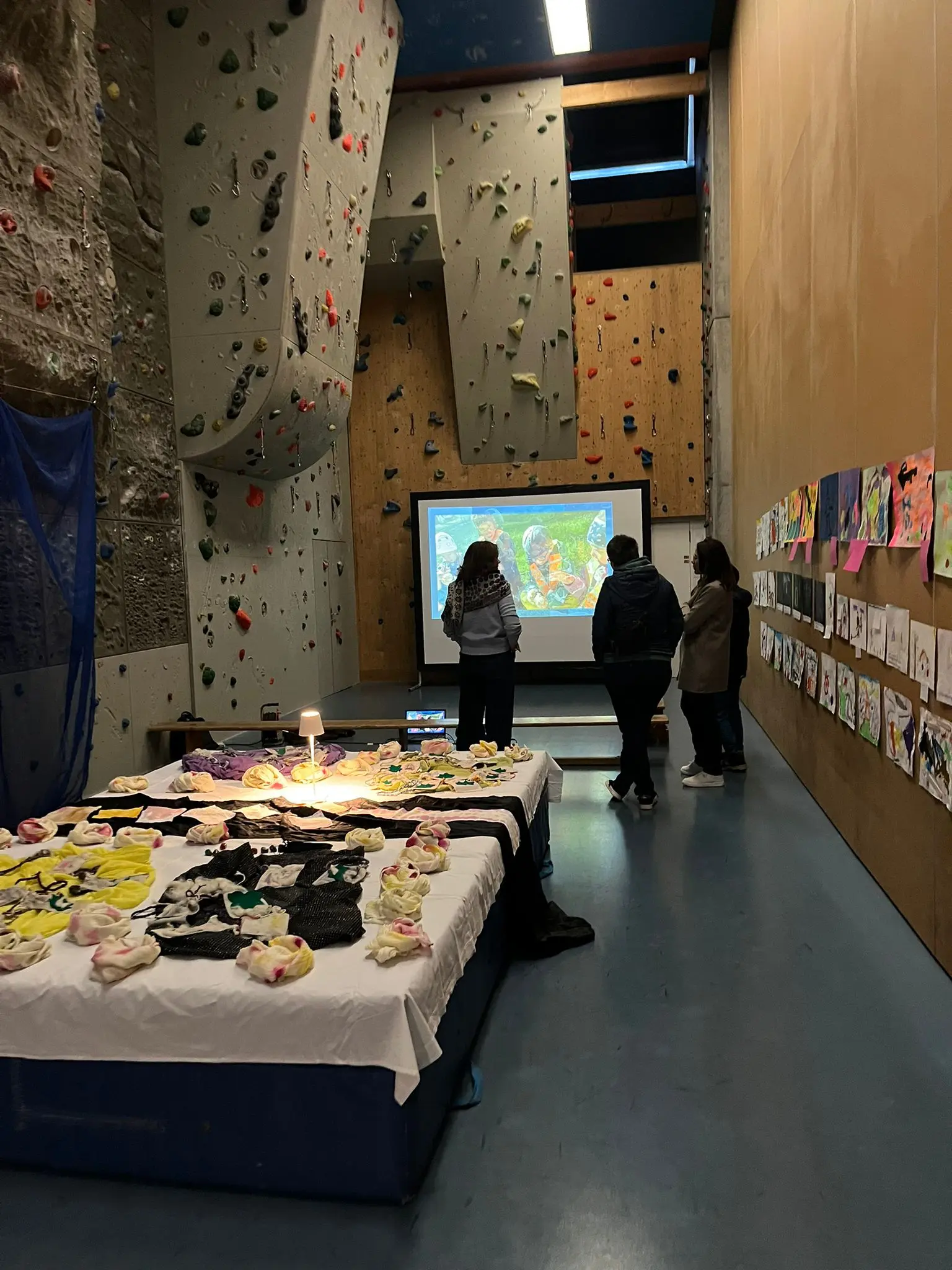 Una sala con pareti per arrampicata, persone in piedi davanti a uno schermo. Su un tavolo ci sono opere d'arte e oggetti decorativi, mentre le pareti sono decorate con disegni.