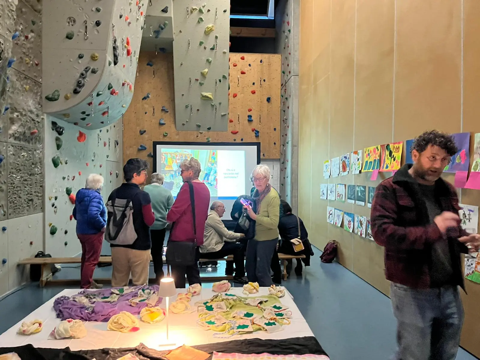 Un gruppo di persone in un ambiente di arrampicata sportiva, con pareti attrezzate per scalatori. Si vedono tavoli con opere d'arte e un proiettore attivo. Un uomo con barba e capelli ricci interagisce con gli altri.