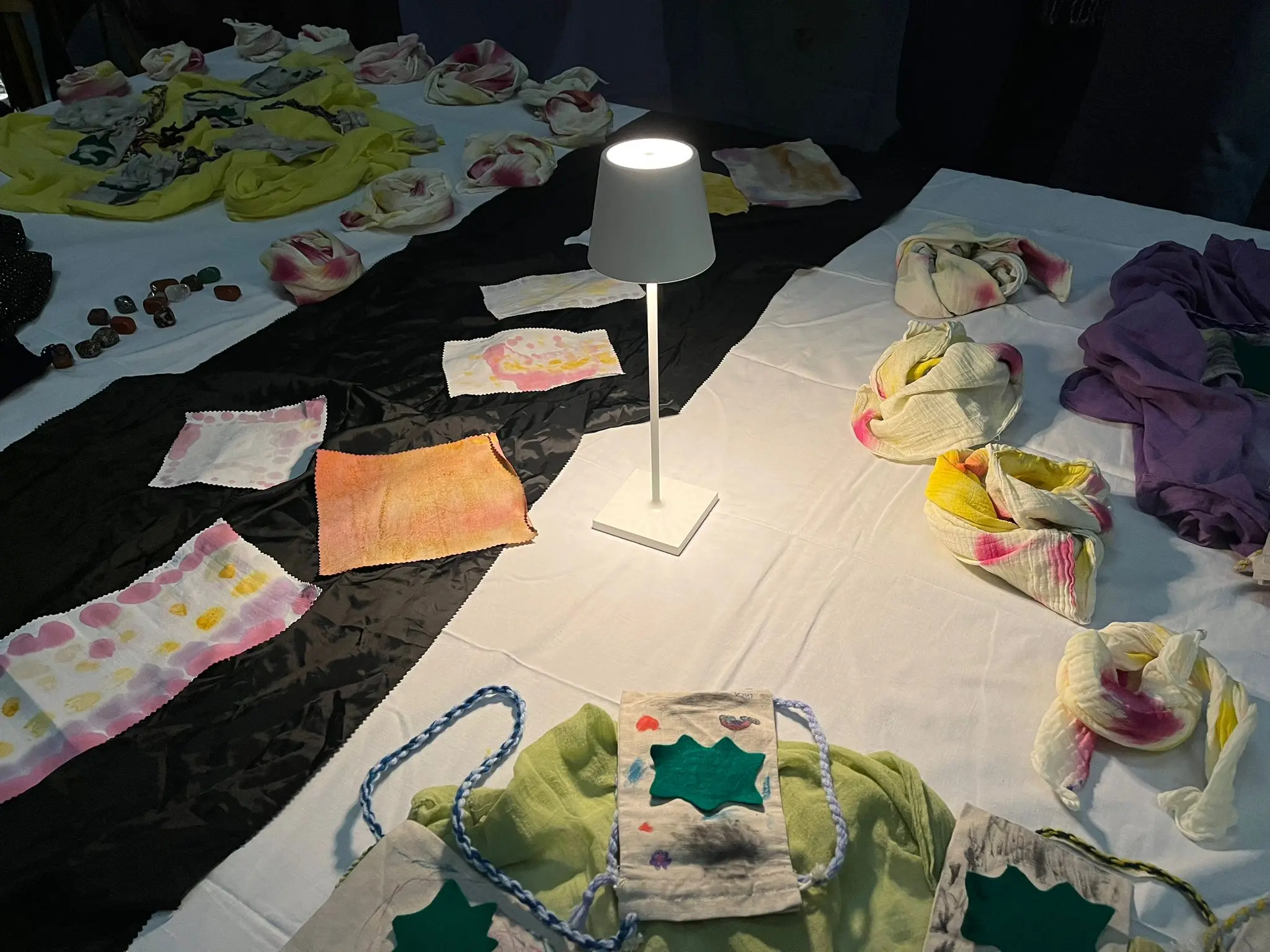 Scialli e tessuti colorati disposti su un tavolo, con un lampada bianca che illumina al centro. Vari motivi e textures visibili.