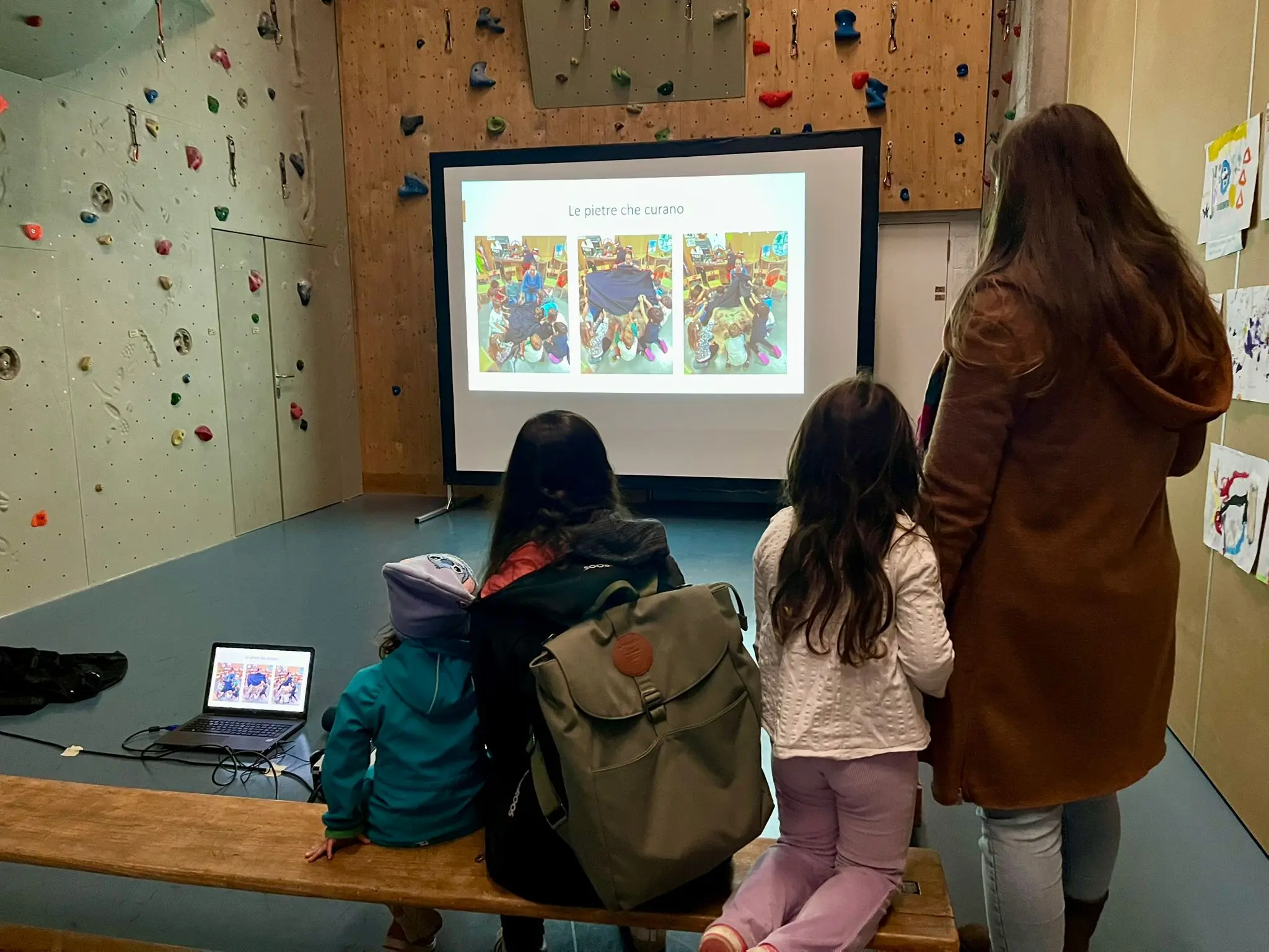 Un gruppo di persone, tra cui due bambini, osserva una presentazione proiettata su uno schermo grande. Sullo sfondo si vede un muro da arrampicata e una panchina di legno.
