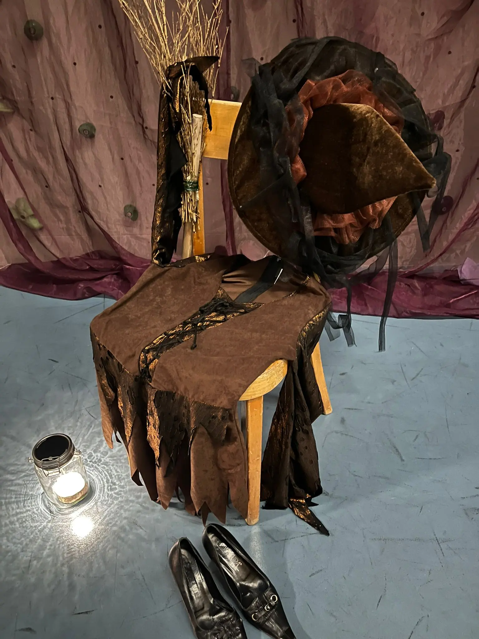 Una sedia decorata con un tessuto marrone scuro e un cappello grande, circondata da rami secchi. Ai piedi ci sono scarpe nere lucide e una lanterna accesa. Lo sfondo è di un colore porpora.