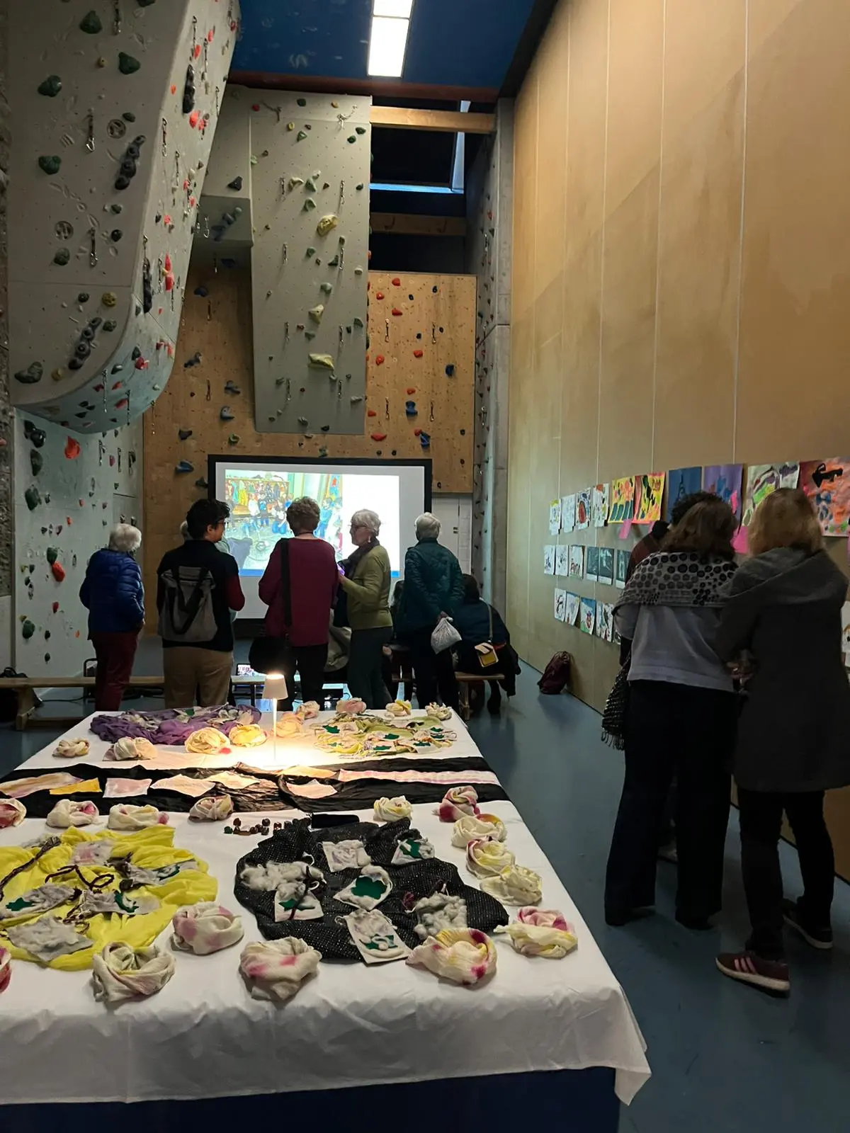 Un gruppo di persone sta osservando un proiettore mentre si trovano in una palestra di arrampicata. In primo piano, un tavolo è decorato con opere artistiche e oggetti vari. Le pareti sono adornate con disegni e dipinti.