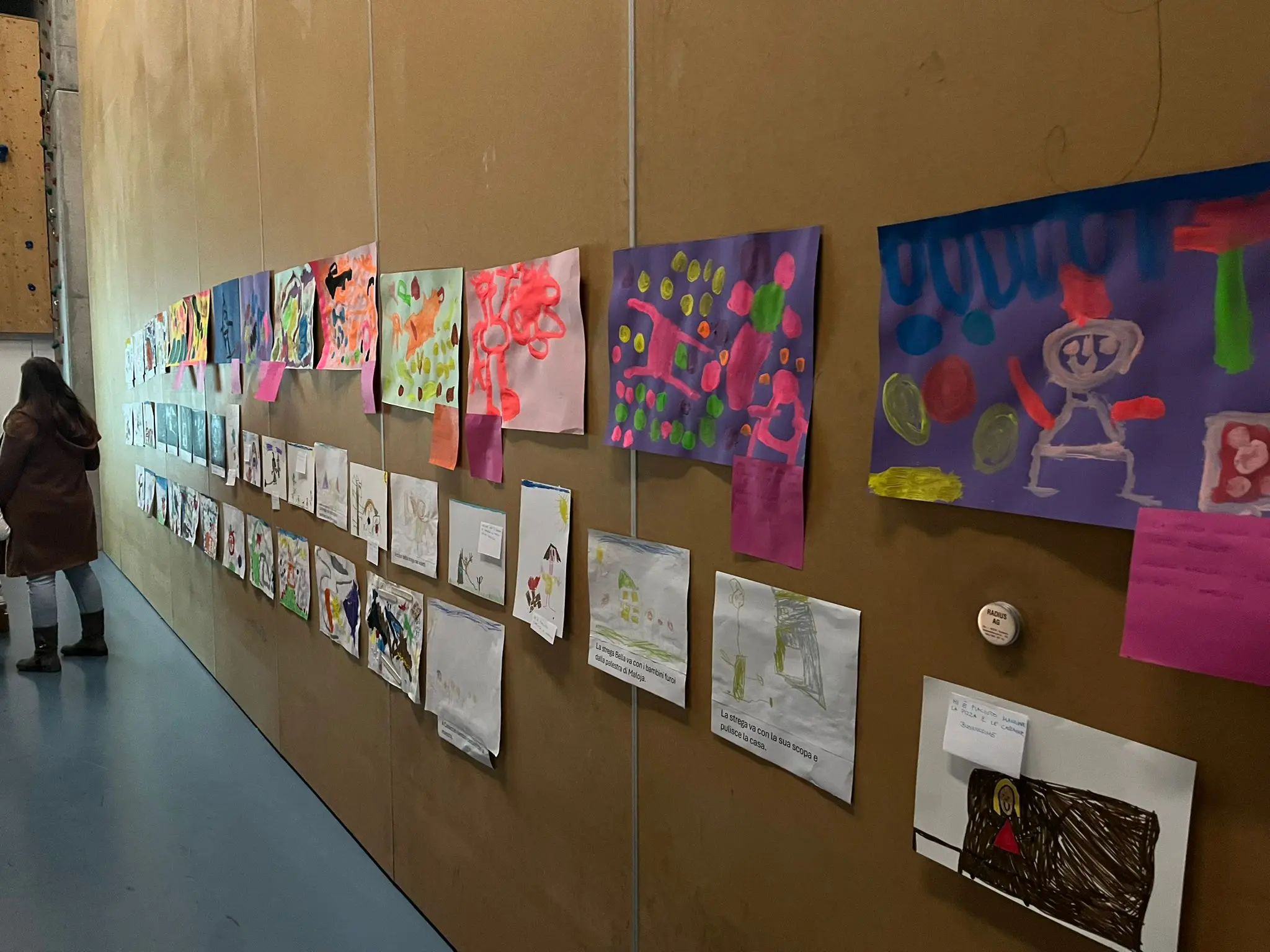 Una parete di esposizione con numerosi disegni colorati, realizzati da bambini, appesi in ordine. Una persona si trova di spalle, osservando i lavori esposti.