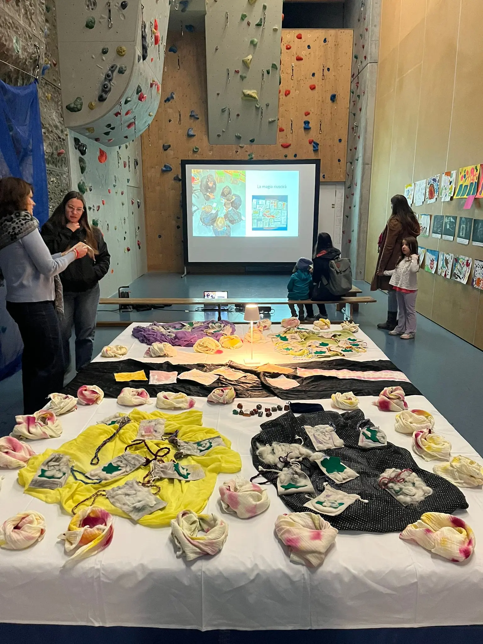 Una grande esposizione di opere d'arte su un tavolo bianco, con maglie colorate e oggetti artistici. Sullo sfondo, un'area di arrampicata e una presentazione proiettata su uno schermo. Persone varie interagiscono e osserv