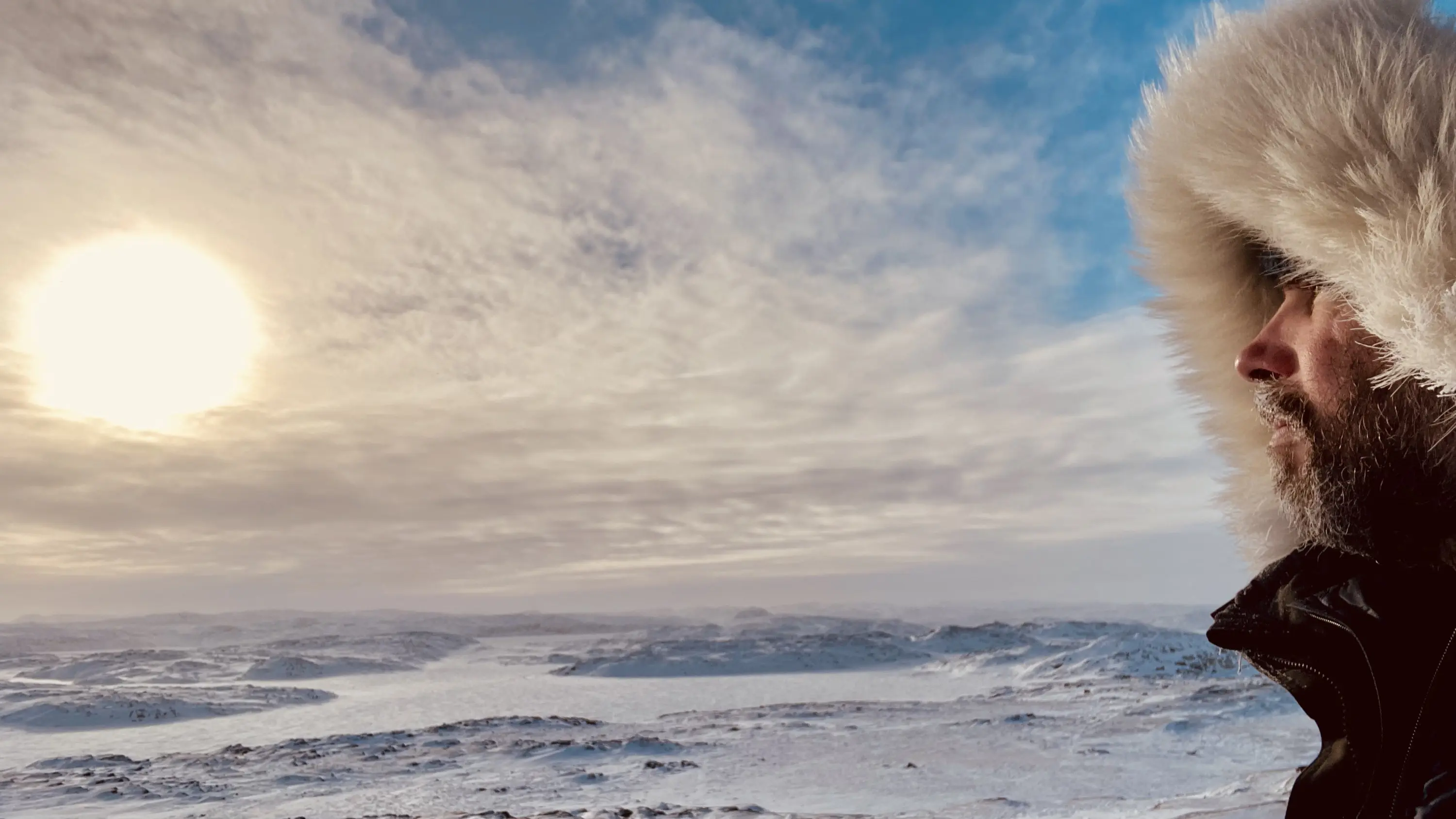 Un uomo con un cappotto di pelliccia osserva il paesaggio innevato e il sole che sorge all'orizzonte, in un ambiente gelido e panoramico.