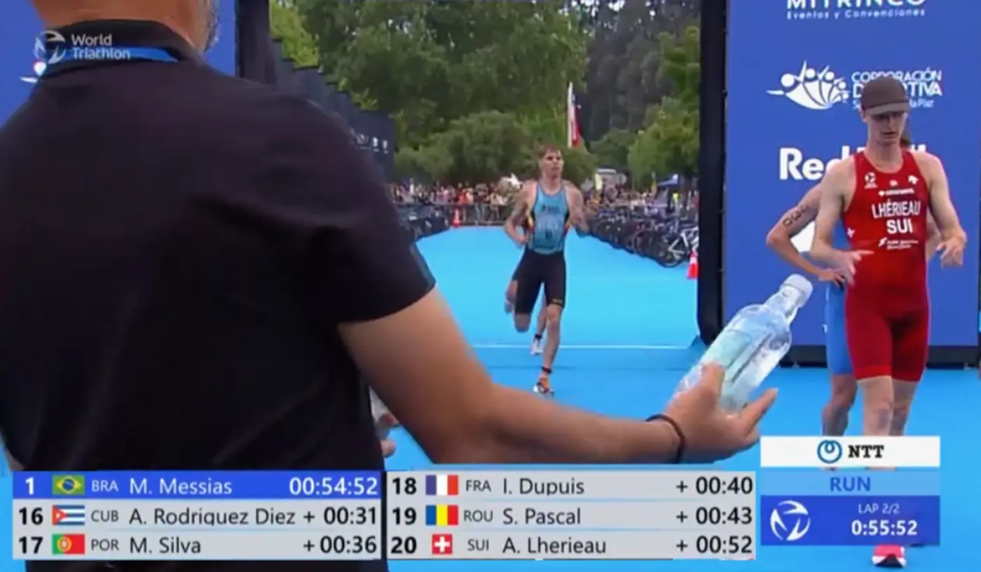 Un triatleta taglia il traguardo durante una gara, mentre un uomo in primo piano porge una bottiglia d'acqua. Sullo schermo si possono vedere i tempi dei concorrenti e le posizioni.