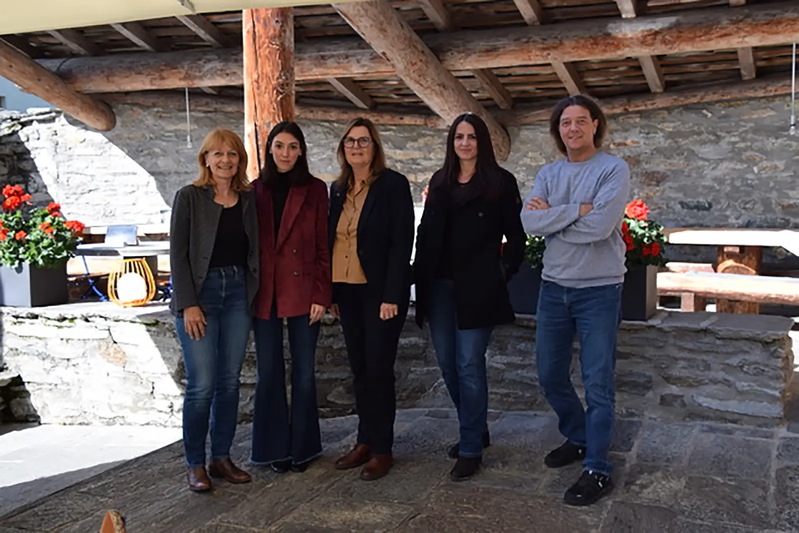 Gruppo di cinque persone in un ambiente rustico e accogliente, con pareti in pietra e piante fiorite sullo sfondo. Tre donne indossano giacche e pantaloni eleganti, mentre una donna è in abbigliamento più casual. Un uomo