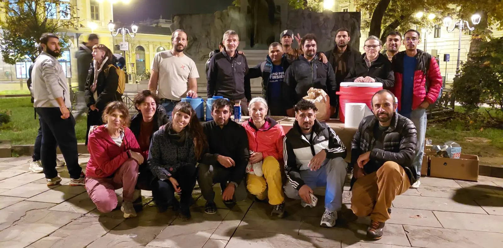 Gruppo di persone sorridenti riunite in un parco di notte, con scatole e attrezzature da campo. Alcuni indossano giacche e abbigliamento sportivo, mentre altri posano in modo informale. Sullo sfondo, illuminazioni urbane