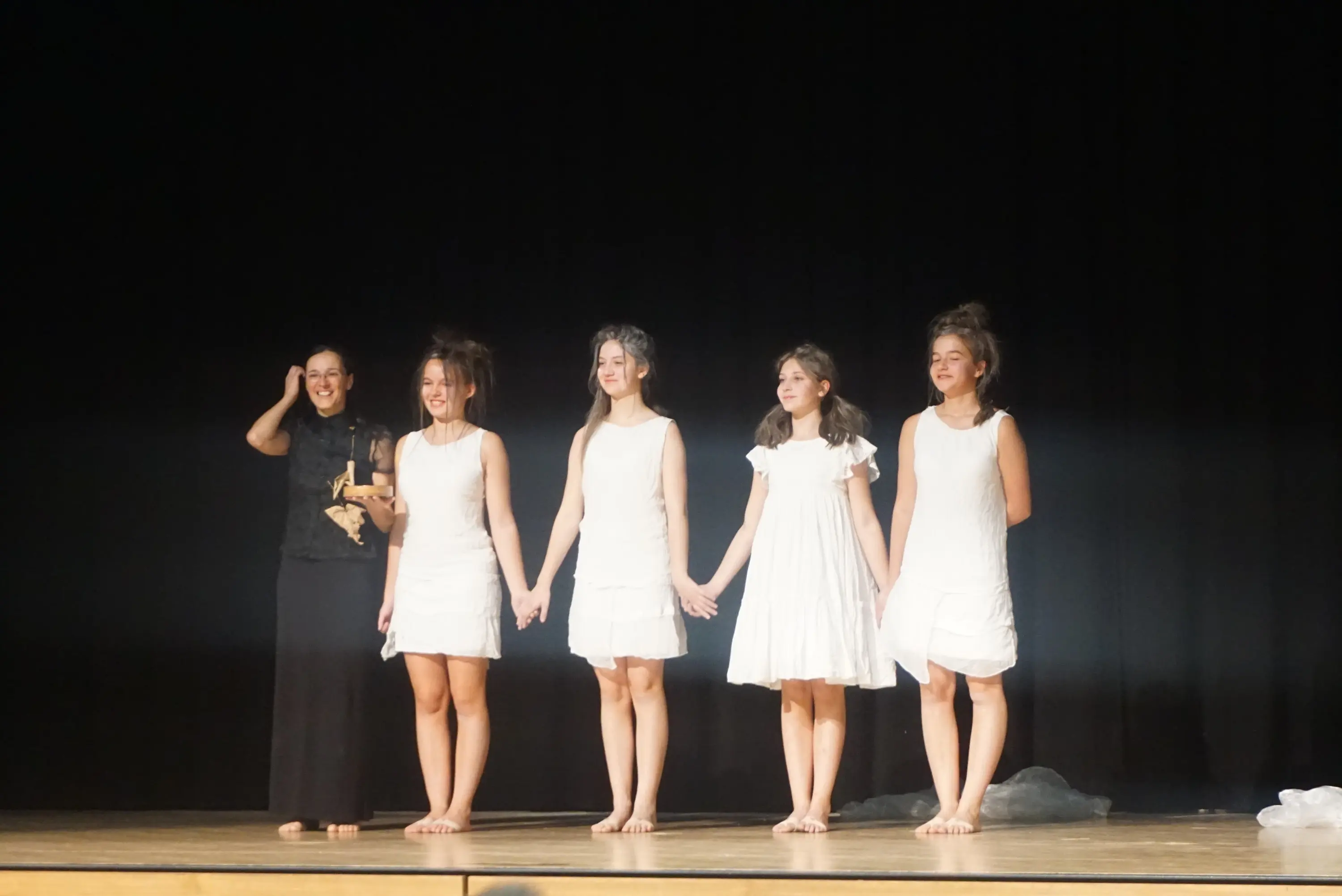 Un gruppo di cinque ragazze, vestite con abiti bianchi, si trovano sul palco. Una di loro, a sinistra, tiene un premio mentre le altre si tengono per mano. Tutte sorridono, mentre il pubblico applaude. Sullo sfondo c'è una