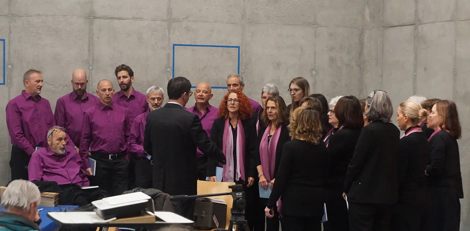 Un coro composto da diverse persone, vestite con camicie e sciarpe di colore viola, canta di fronte a un direttore. Alcuni membri sono seduti, mentre altri si trovano in piedi. Lo sfondo è una parete di cemento.