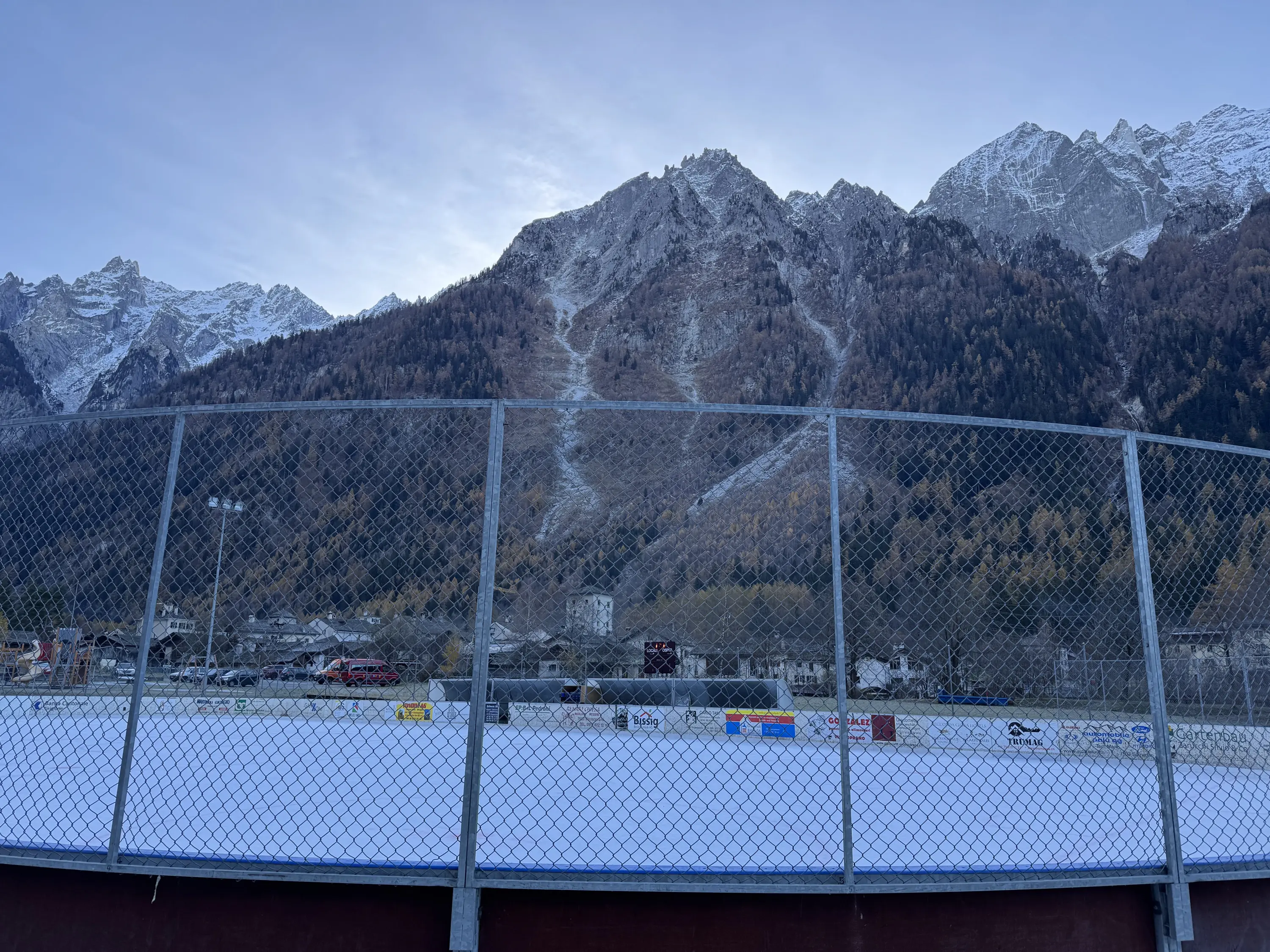 Una pista di ghiaccio all'aperto circondata da montagne innevate e alberi autunnali. Il cielo è sereno e luminoso, mentre una rete metallica delimita l'area di gioco.