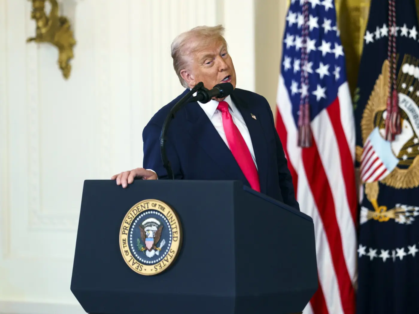 Trump parla a un podio con il sigillo presidenziale, circondato da bandiere americane.