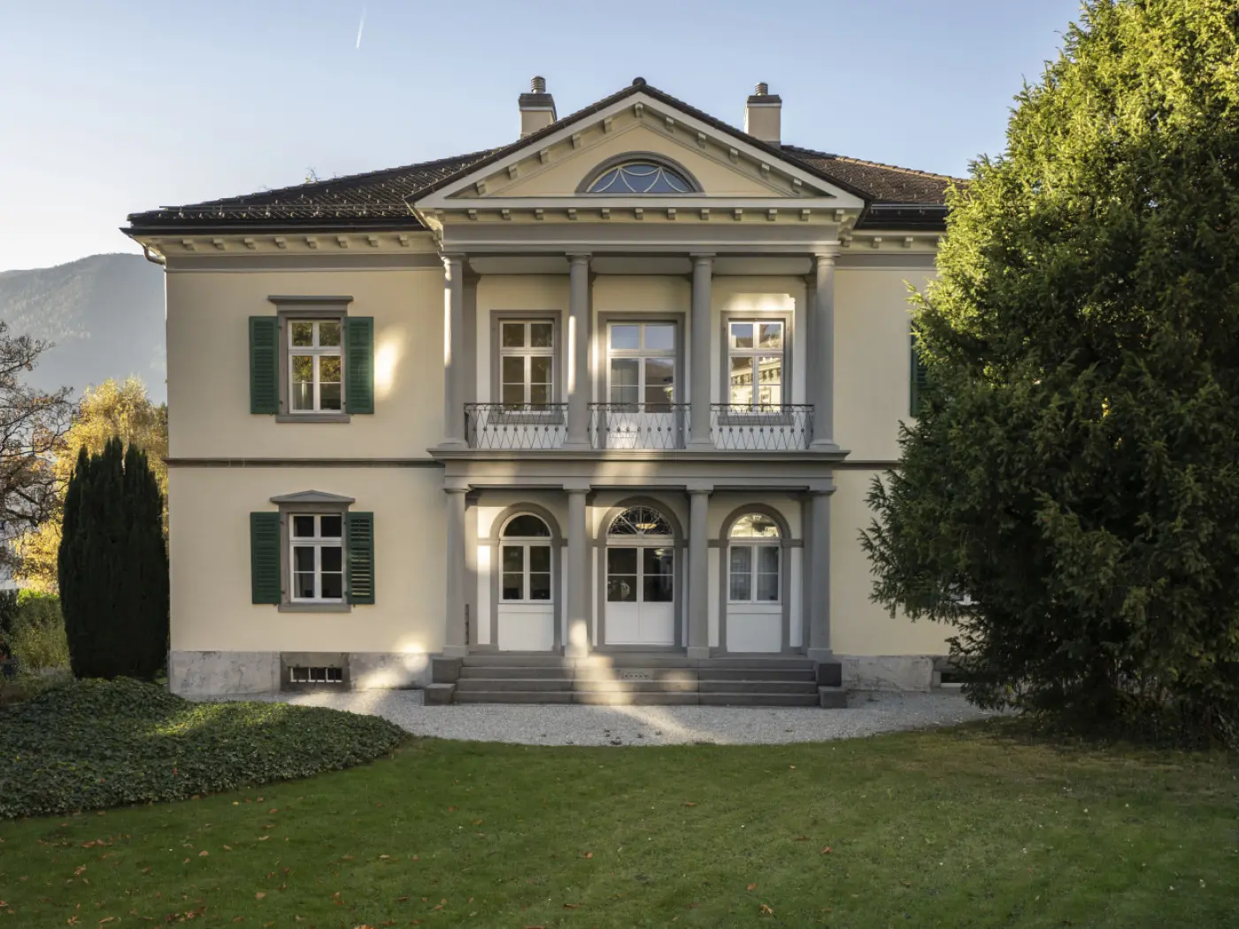 Villa elegante con facciata gialla, colonne bianche e finestre con persiane verdi. Circondata da un giardino curato e alberi.