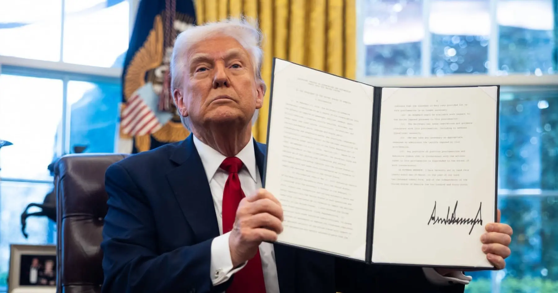 Un uomo in giacca e cravatta rossa tiene in mano un documento firmato, seduto in una stanza con tende gialle e simboli presidenziali.