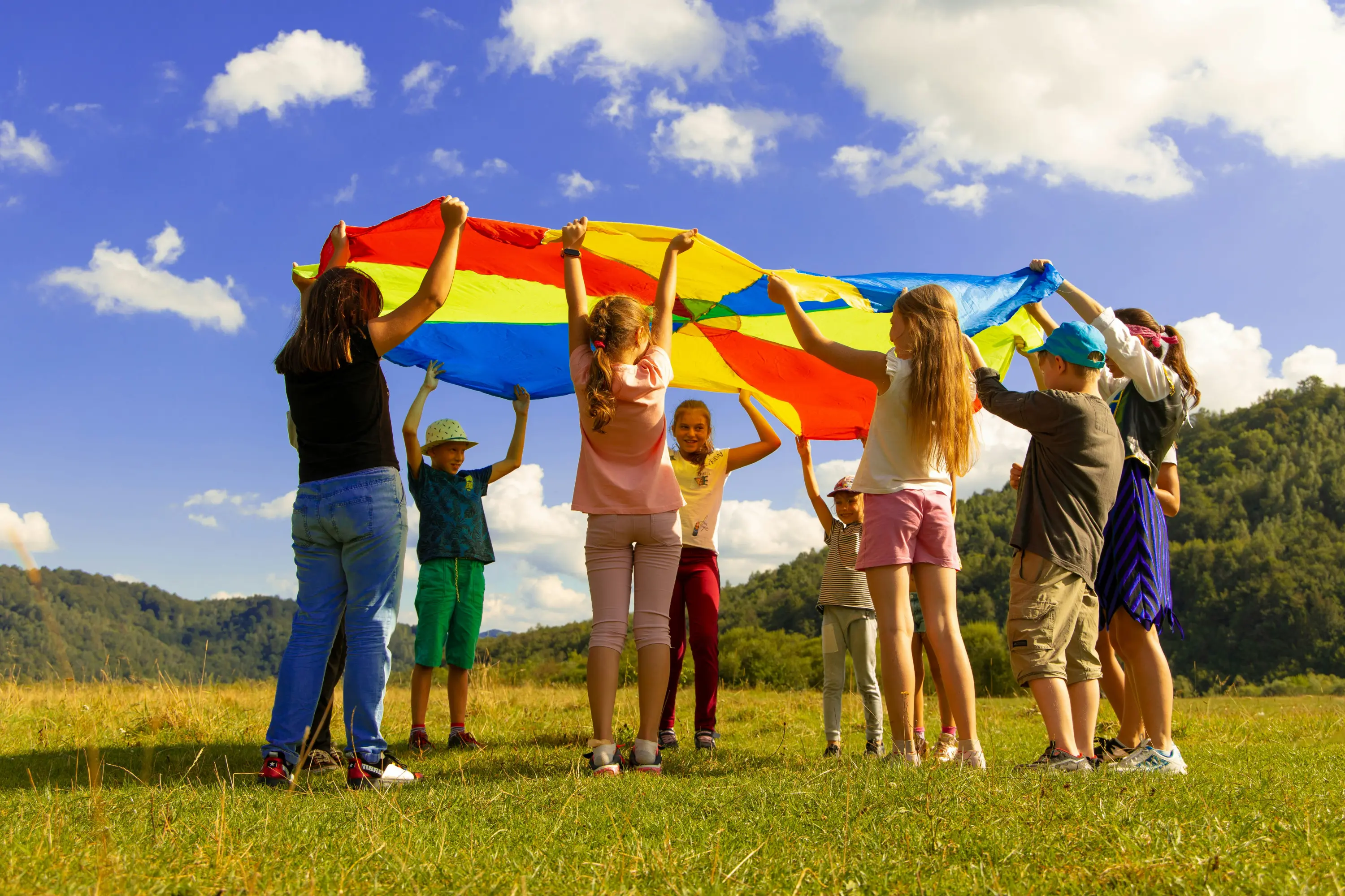 Gruppo di bambini che tengono in alto una grande bandiera colorata in un campo aperto. Sullo sfondo, colline verdi e un cielo blu con nuvole bianche. I bambini sorridono e sembrano divertirsi.