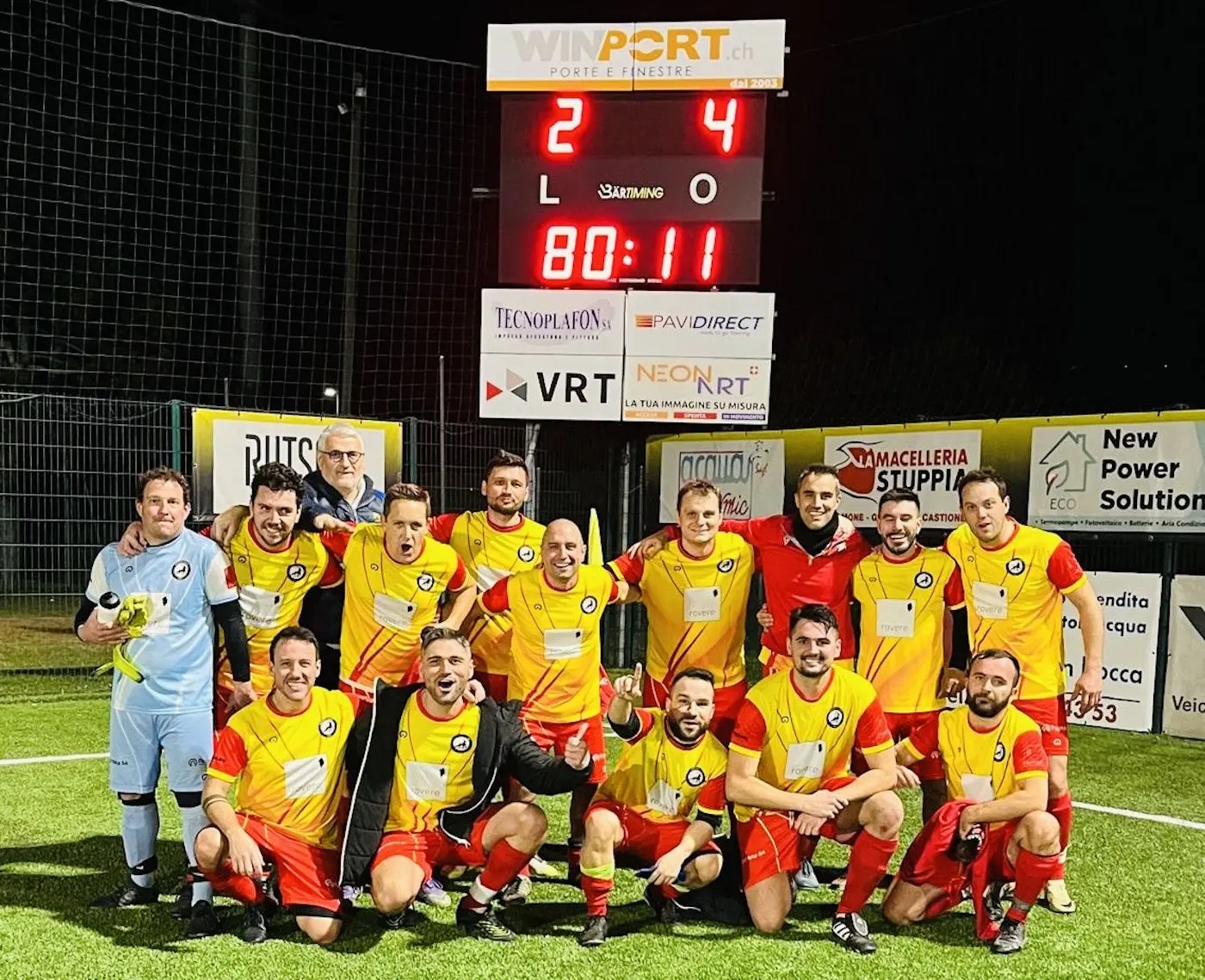 Una squadra di calcio sorride insieme sul campo, indossando maglie arancioni e nere. Sono in posa per una foto di gruppo, con alcuni giocatori in ginocchio e altri in piedi, mentre il punteggio sullo sfondo indica 2-4.
