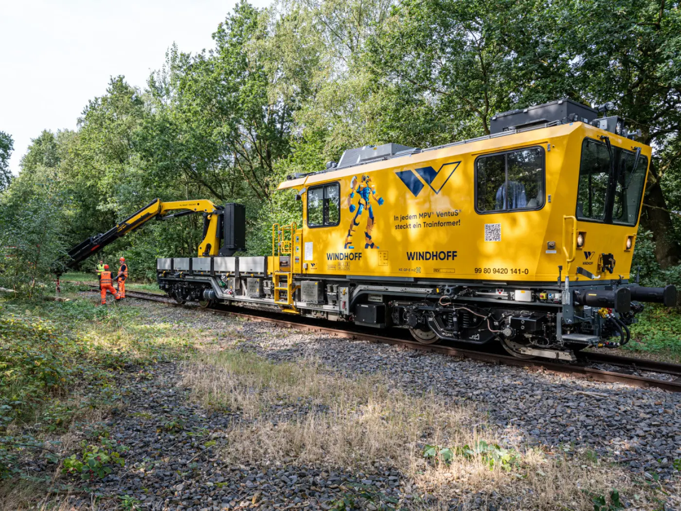 Un veicolo ferroviario giallo dotato di un braccio meccanico stilizzato, parcheggiato su rotaie in un'area alberata. Due lavoratori in tuta arancione stanno esaminando il macchinario.