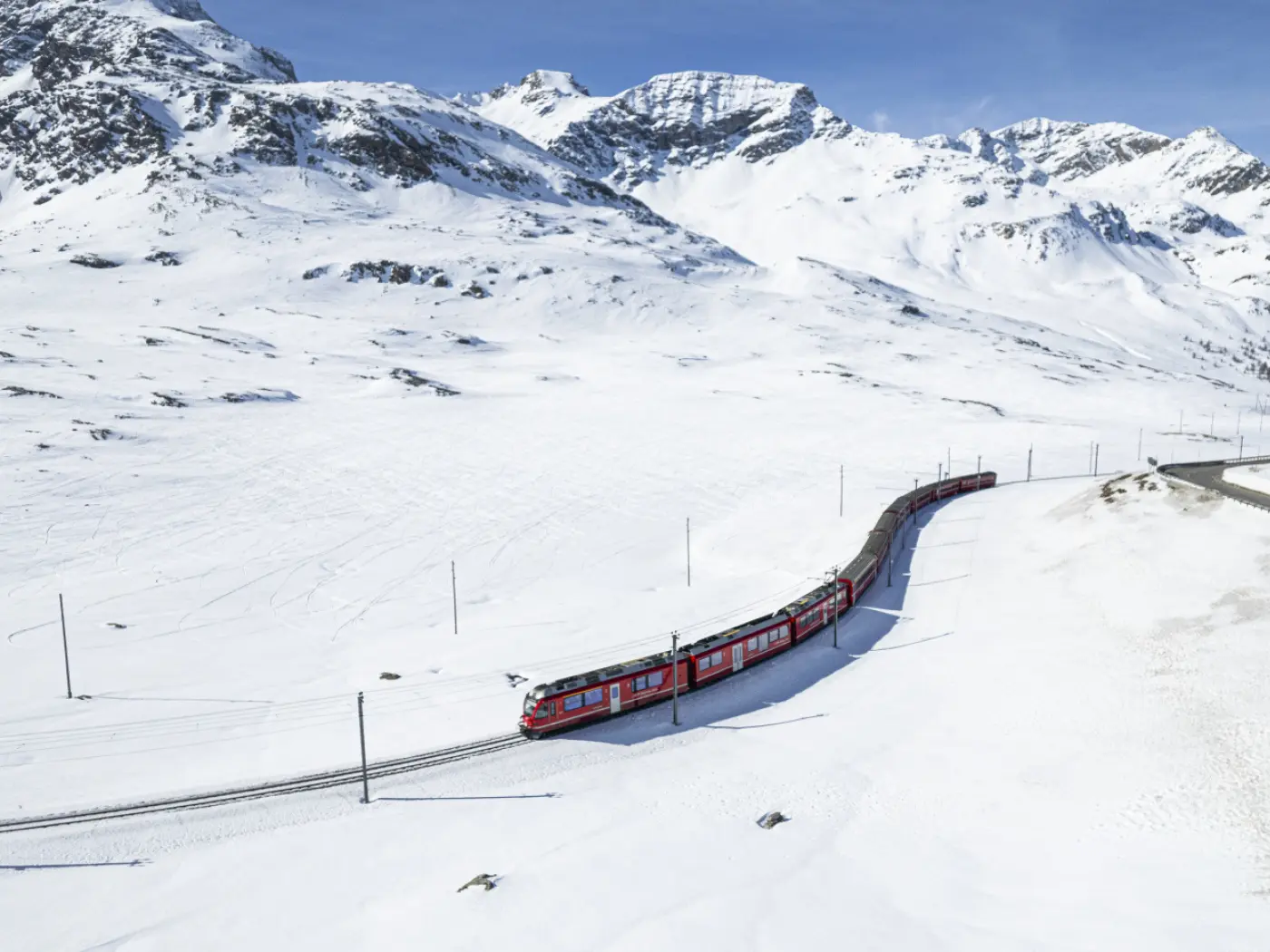 Un treno rosso curva attraverso un paesaggio innevato, circondato da alte montagne. Il cielo è sereno e blu.