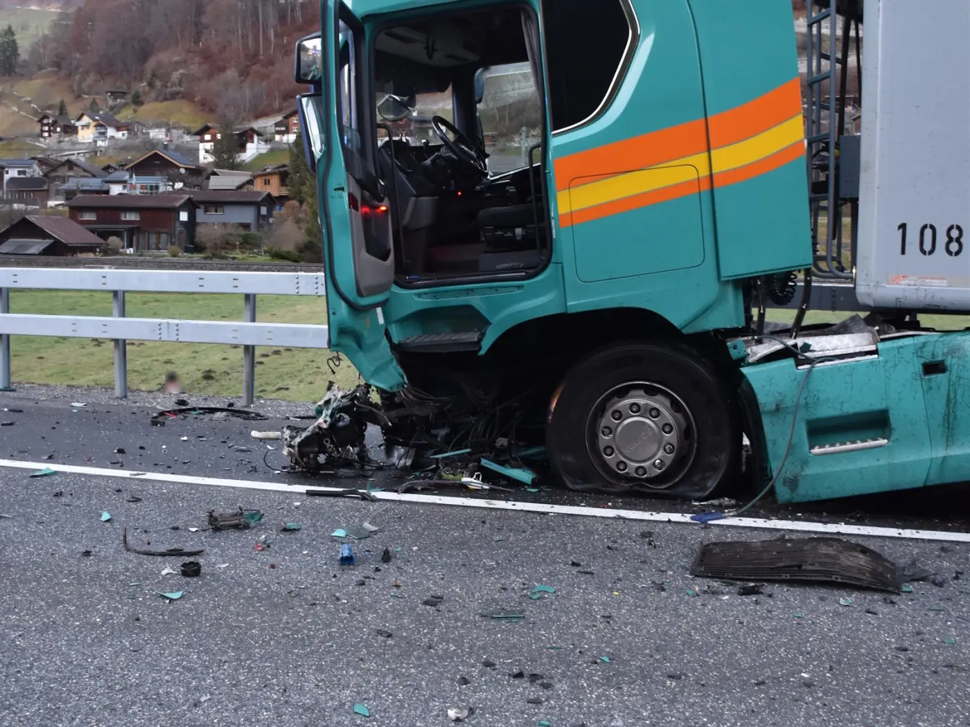 Camion coinvolto in un incidente stradale, con la parte anteriore danneggiata e detriti sparsi sulla carreggiata. Sullo sfondo, visibili case e un paesaggio montano.