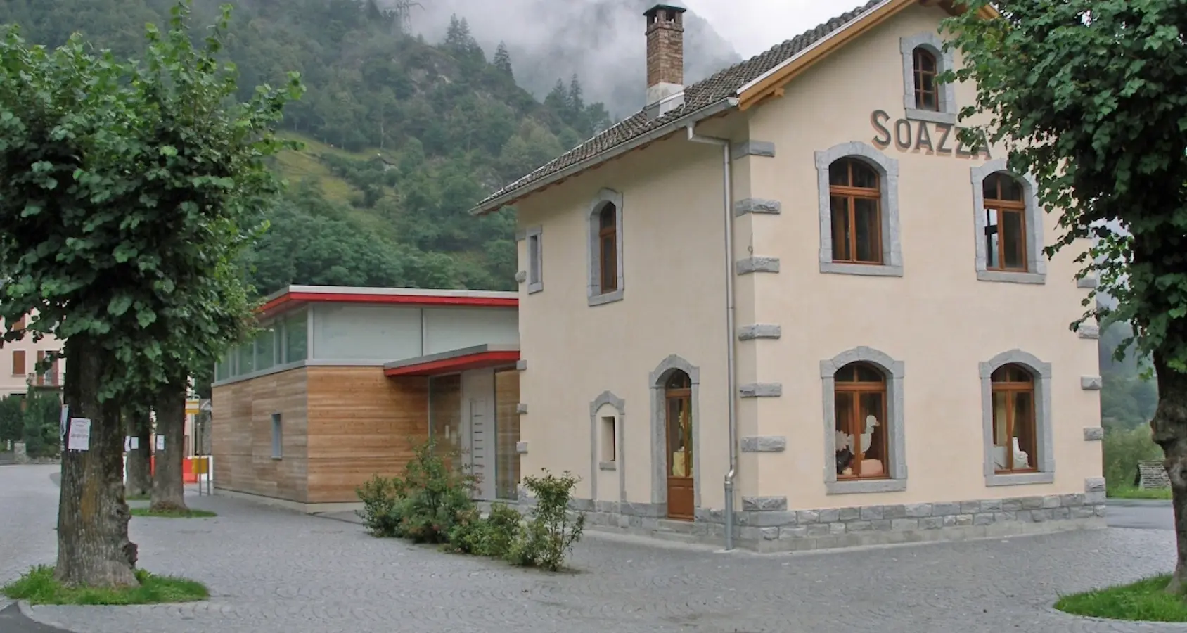 Un edificio a due piani con pareti di un beige chiaro e finestre con cornici grigie. Sul lato sinistro, una struttura moderna in legno e vetro si collega al vecchio edificio. Sullo sfondo, colline verdi e nuvole basse.