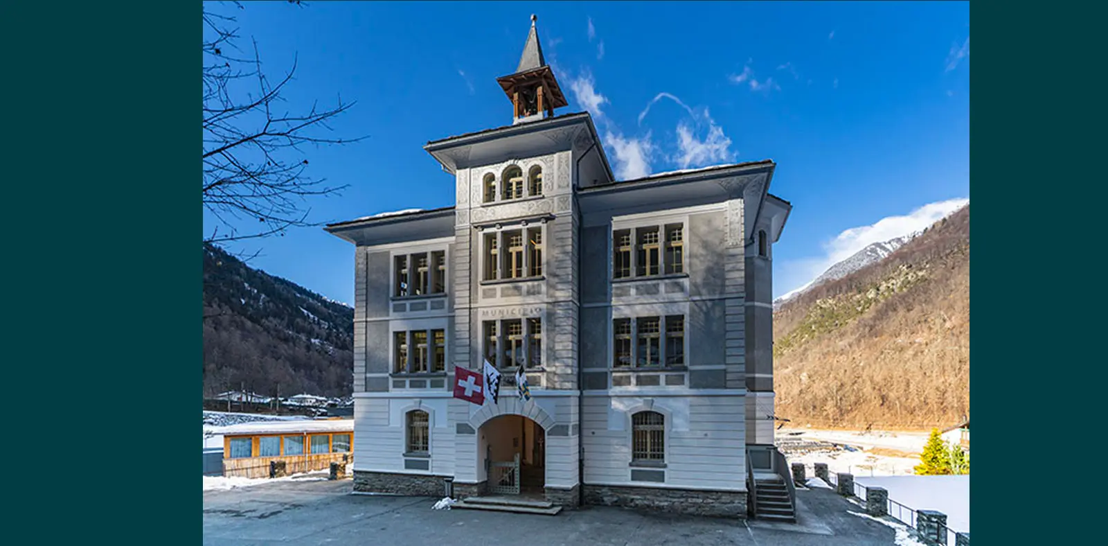 Edificio storico a tre piani con una torre, caratterizzato da una facciata bianca e grigia. La bandiera svizzera sventola all'ingresso, circondato da un paesaggio montano coperto di neve. Cielo azzurro con alcune nu