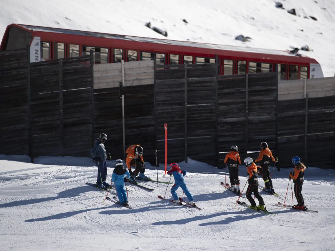 Un gruppo di bambini in tuta da sci, accompagnati da istruttori, scia su una pista innevata accanto a una costruzione di legno e a una funivia rossa.